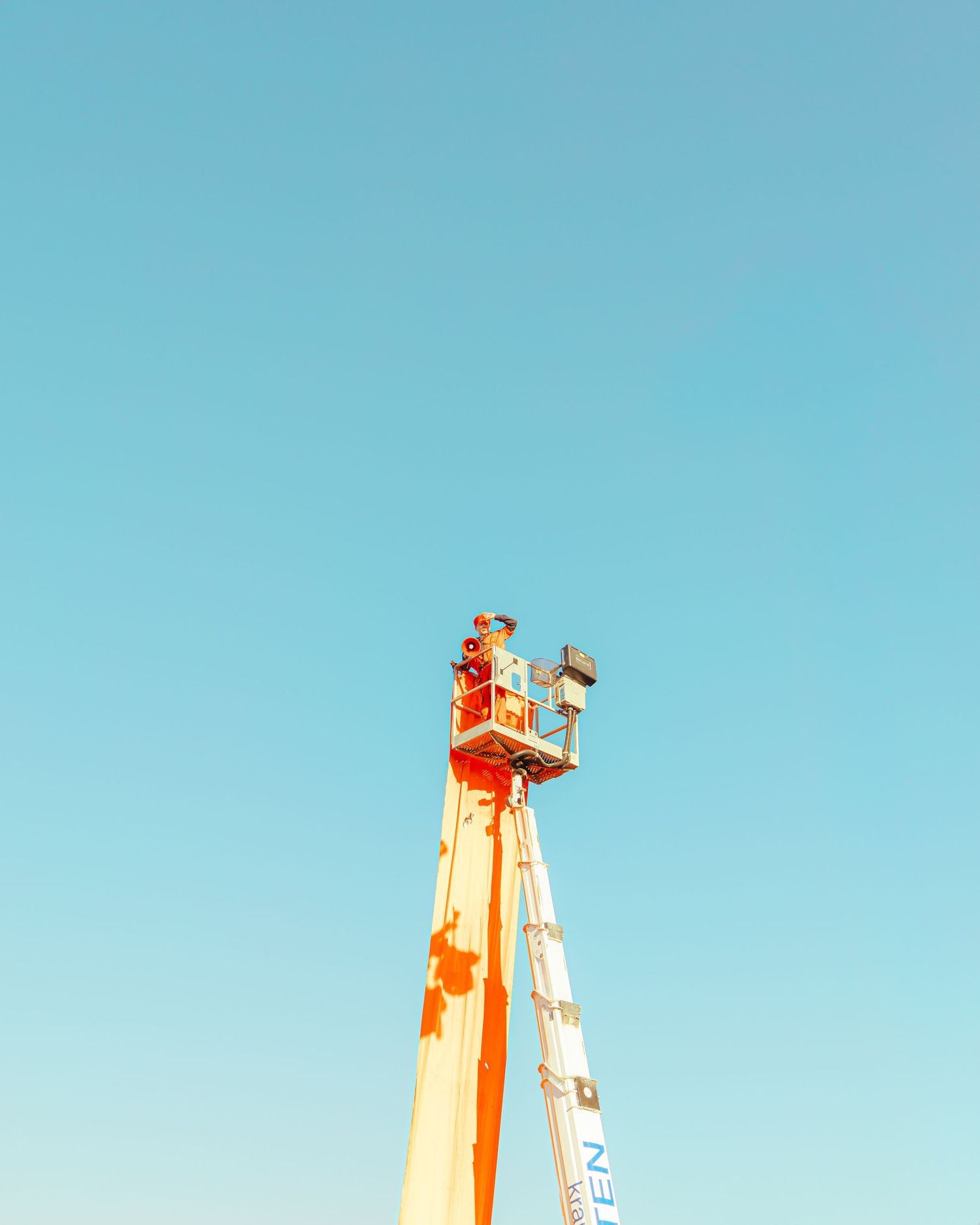 Orange aerial work platform lift extended against a clear blue sky on a sunny day.