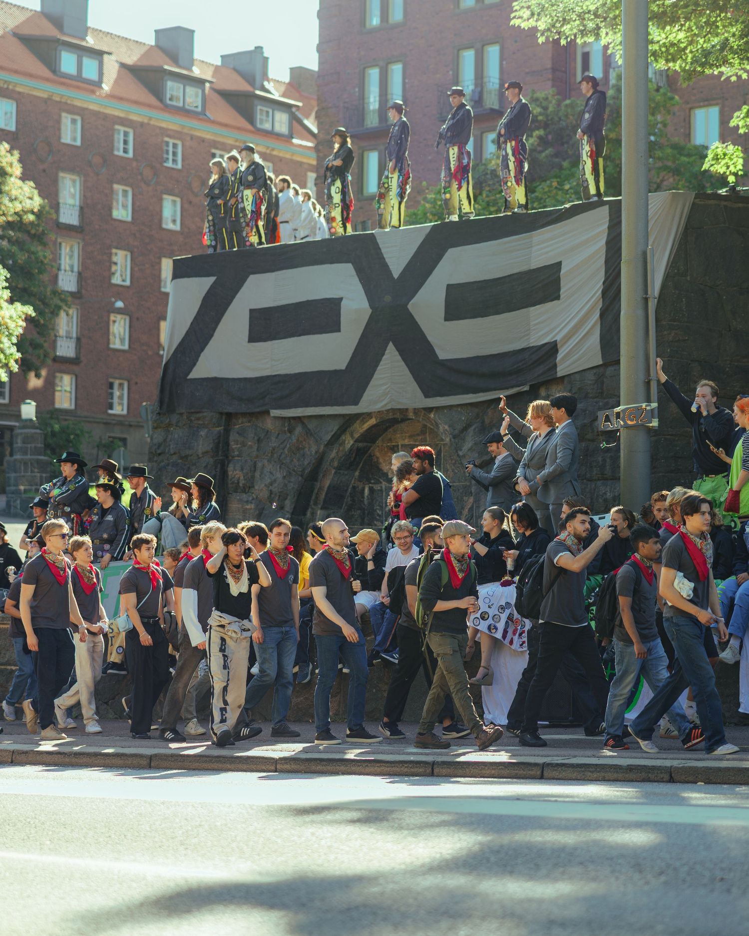 Large crowd marching in street past bold black banner with logo, urban buildings visible in sunny background.