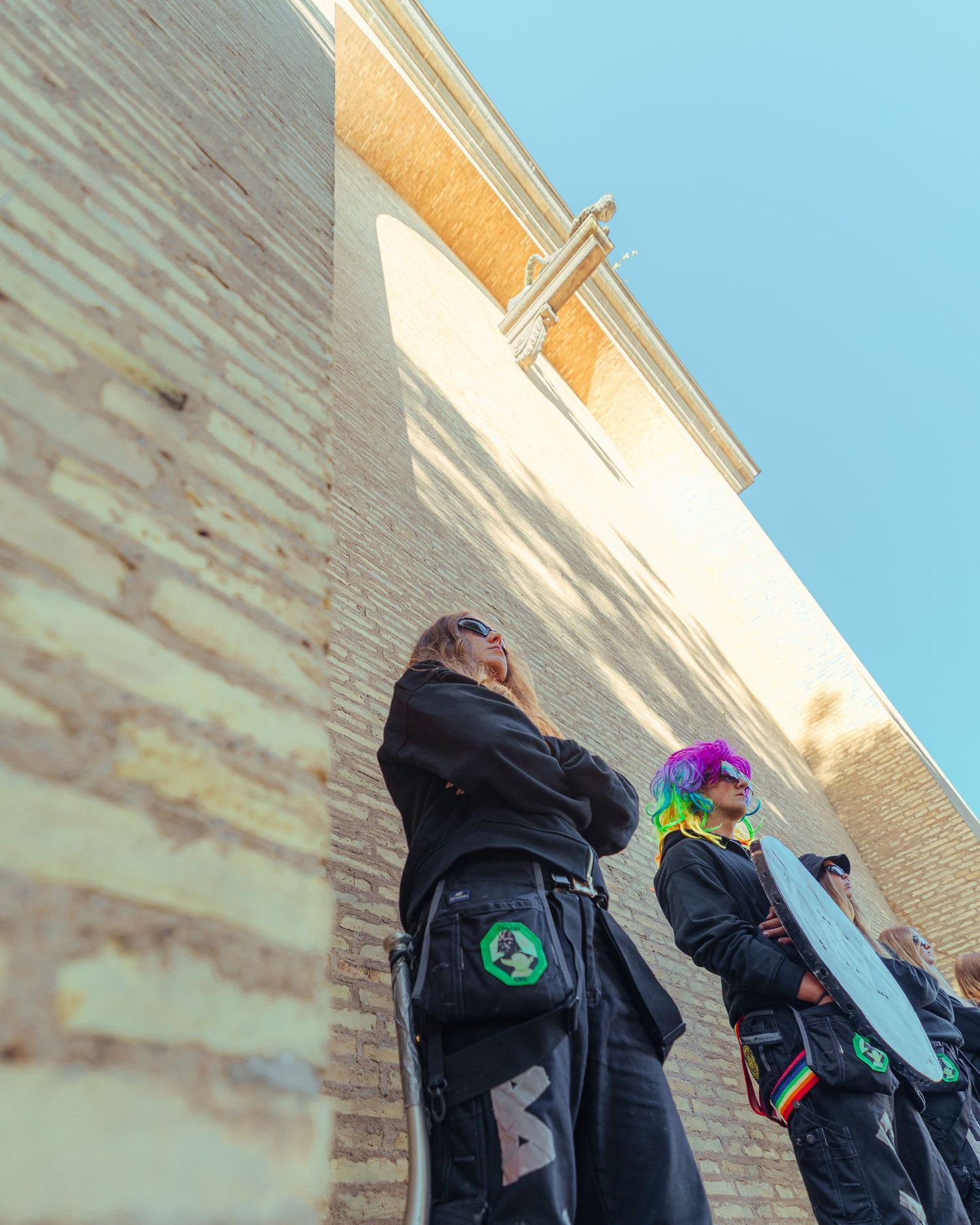 Two people in black outfits with colorful accessories stand against a brick wall under a blue sky.