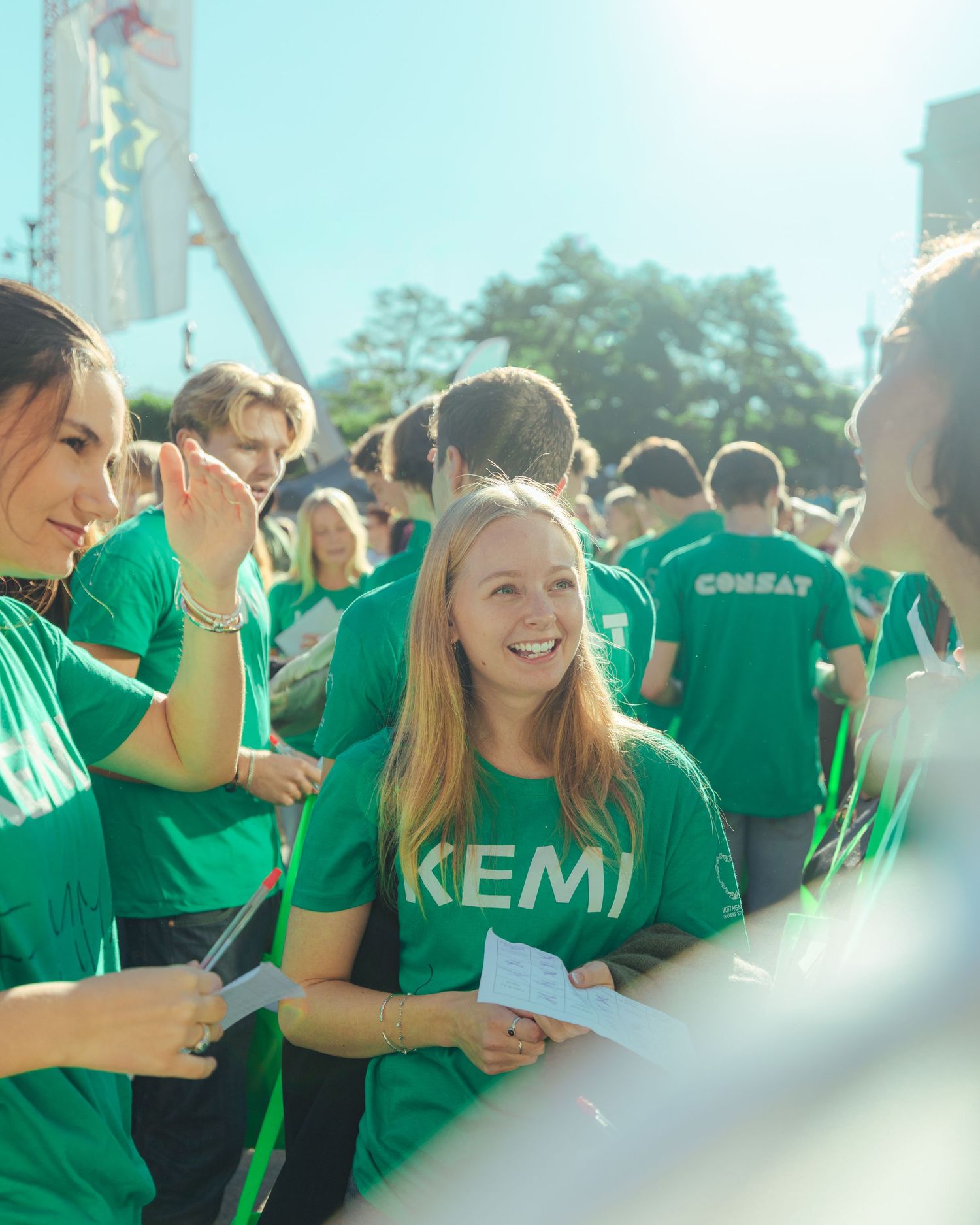 Students in green KEMI t-shirts smile and socialize outdoors at a sunny university orientation event.