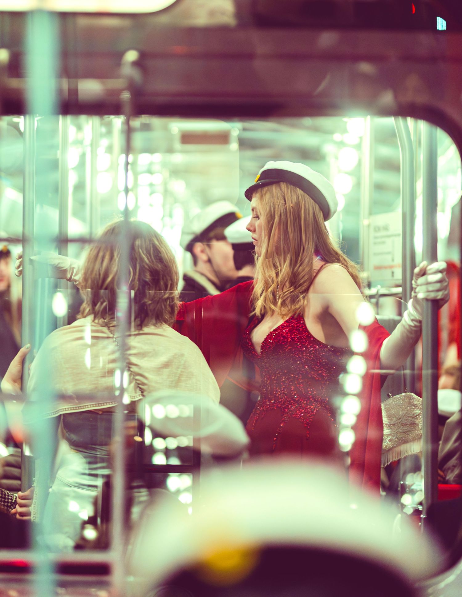 Elegant woman in red sequin dress and white hat standing in a brightly lit tram.