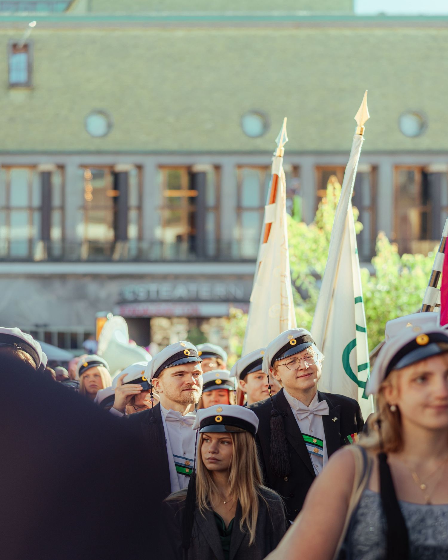 Students in white graduation caps carry flags during an outdoor ceremony near a historic building.