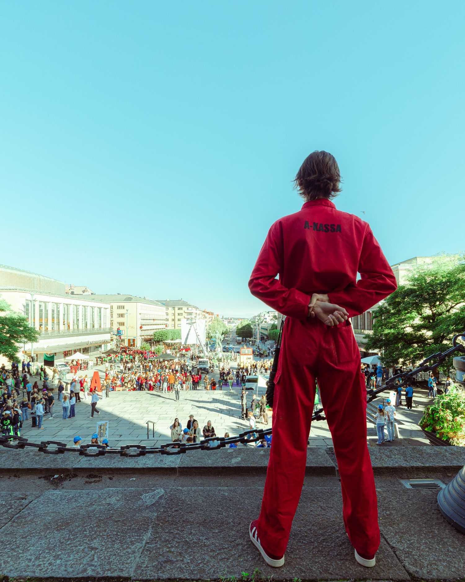 Person in red jumpsuit stands on elevated platform overlooking a crowded city square on a sunny day.