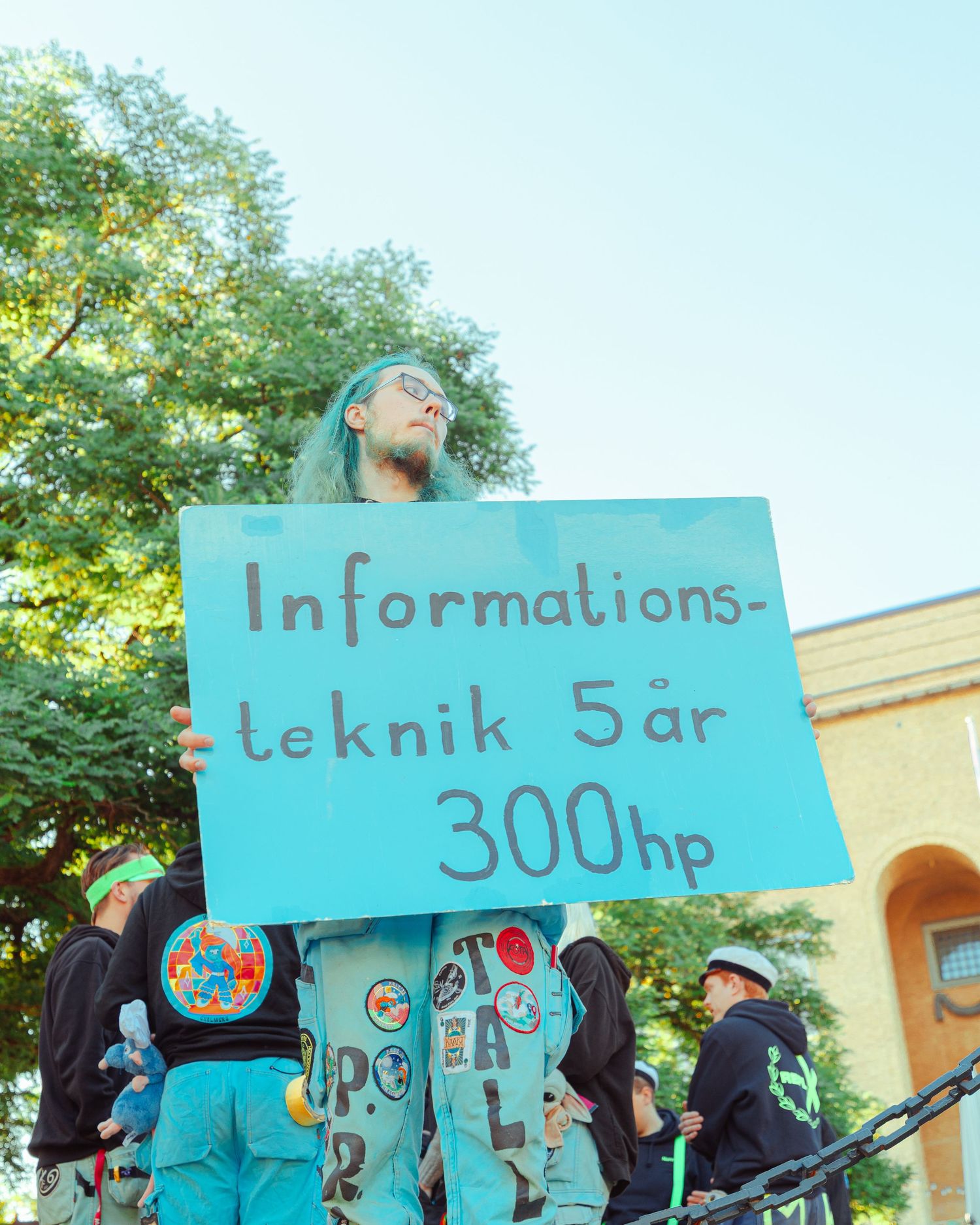 Person holding a blue sign reading Informationsteknik 5 år 300hp at an outdoor student graduation celebration.