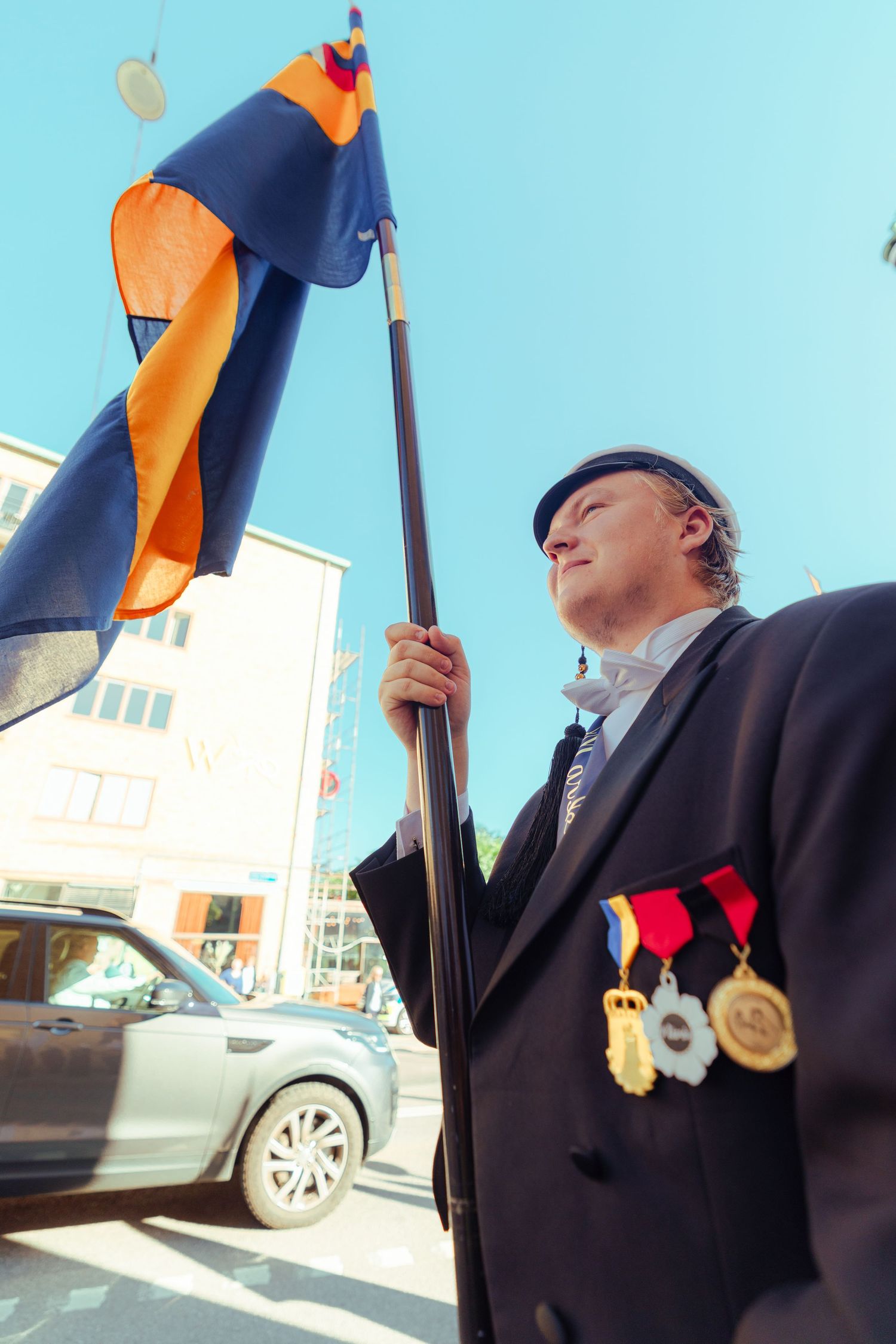 Man in formal attire holding a flag and wearing medals at an outdoor ceremony on a sunny day.