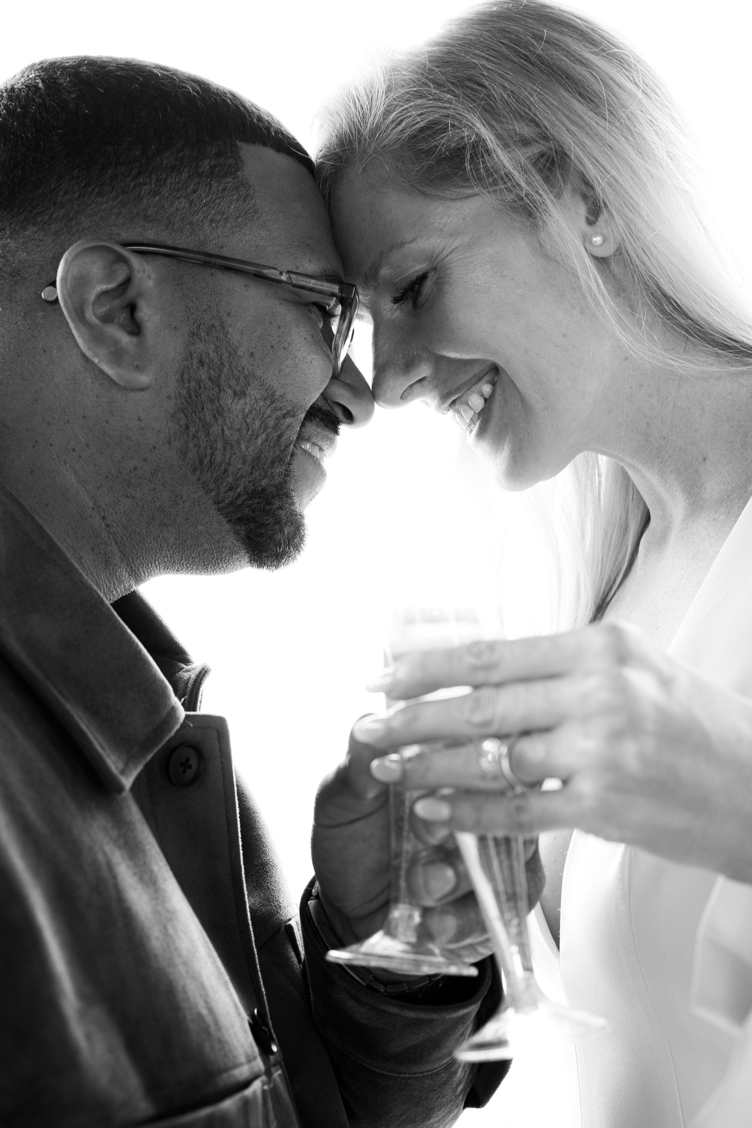 Black and white photo of a smiling couple touching foreheads and clinking champagne glasses in a romantic moment.