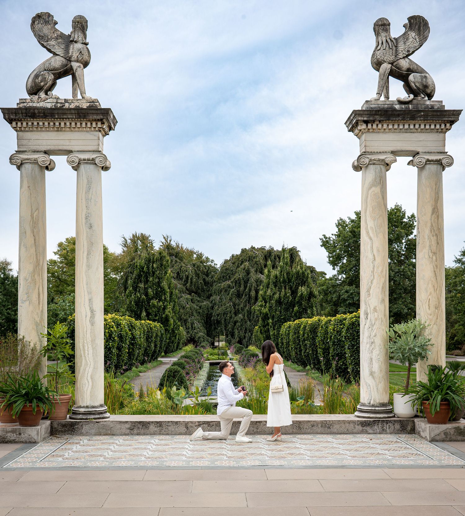 Man proposes to woman on one knee between ornate columns with griffin statues in a formal garden setting.