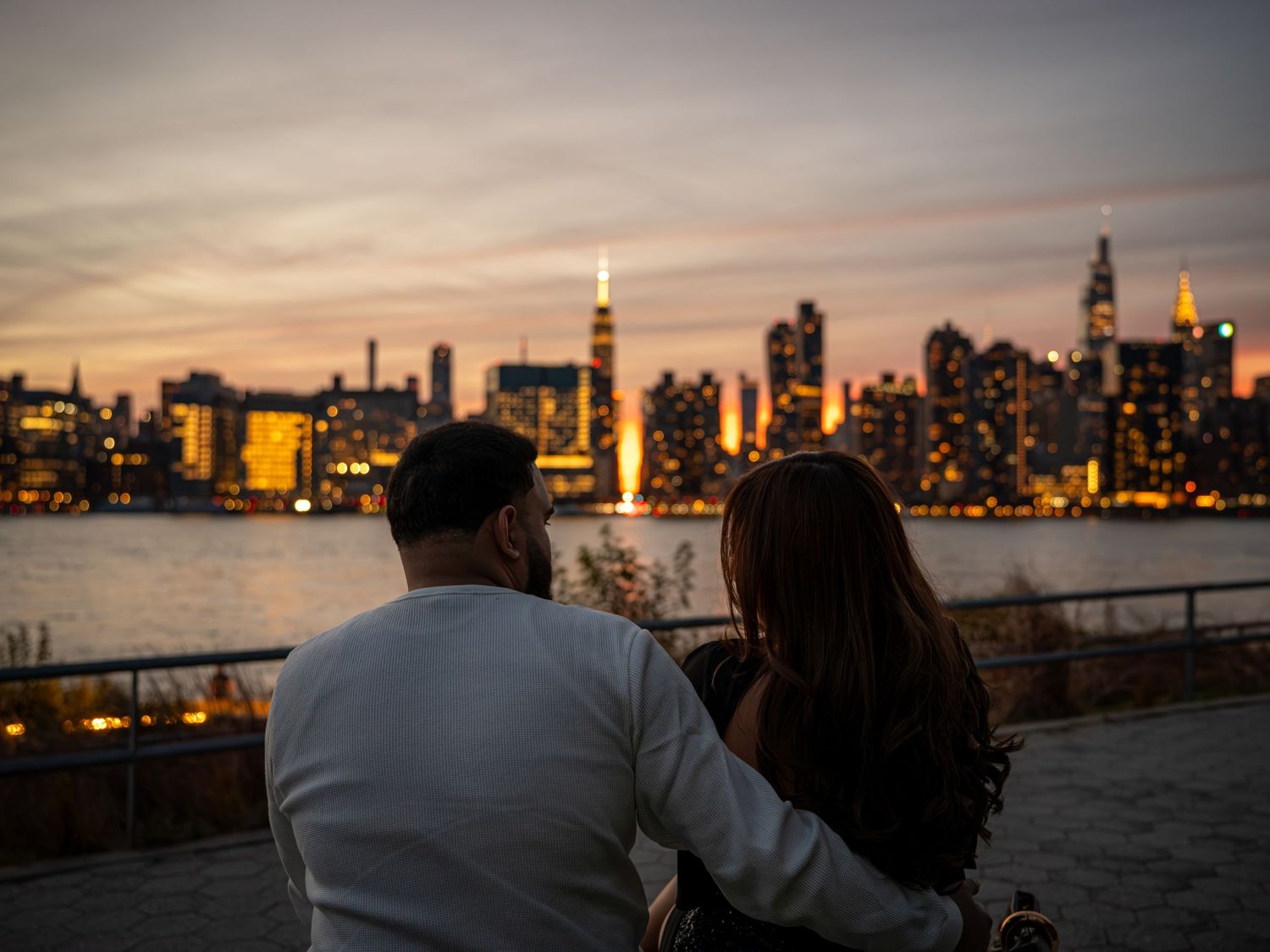Couple sitting together enjoying a romantic NYC skyline sunset view across the water with illuminated skyscrapers.