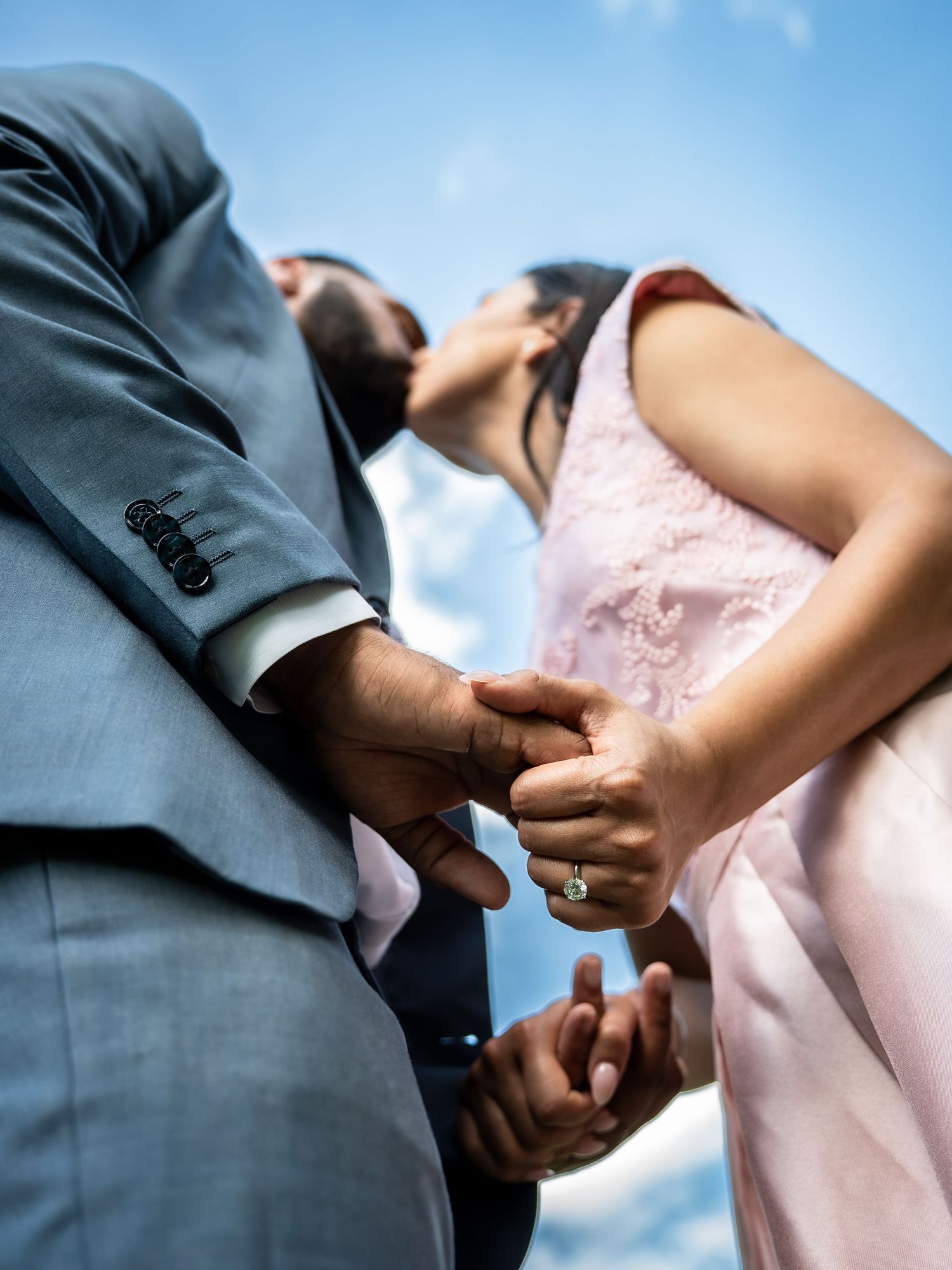 Couple holding hands during outdoor wedding ceremony, bride wearing pink lace dress and engagement ring, groom in gray suit.
