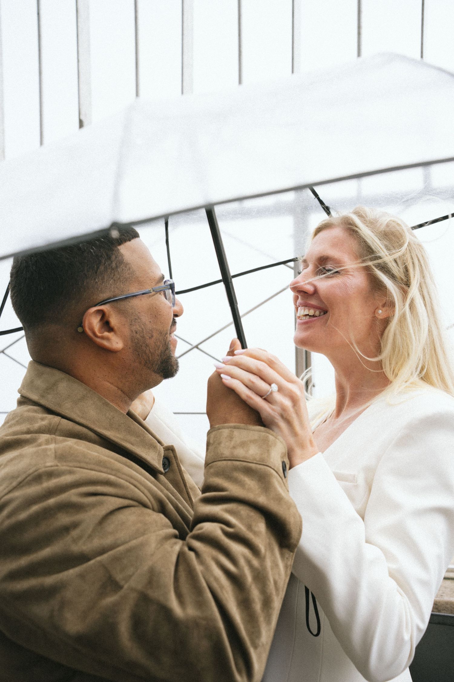 Interracial couple smiling at each other under a white umbrella, woman in white blazer holding hands with man in tan jacket.