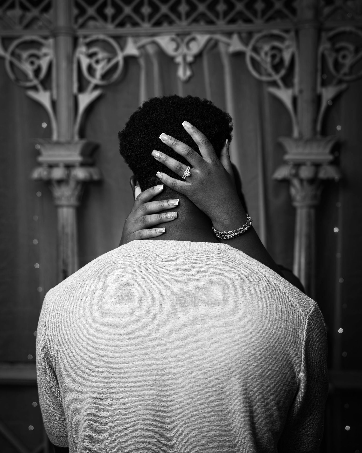 Black and white photo of a person seen from behind, hand resting on neck, wearing a sweater near ornate architecture.