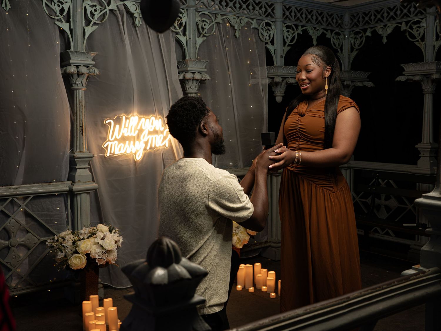 Man proposes to woman on one knee surrounded by candles and a neon sign reading Will You Marry Me.