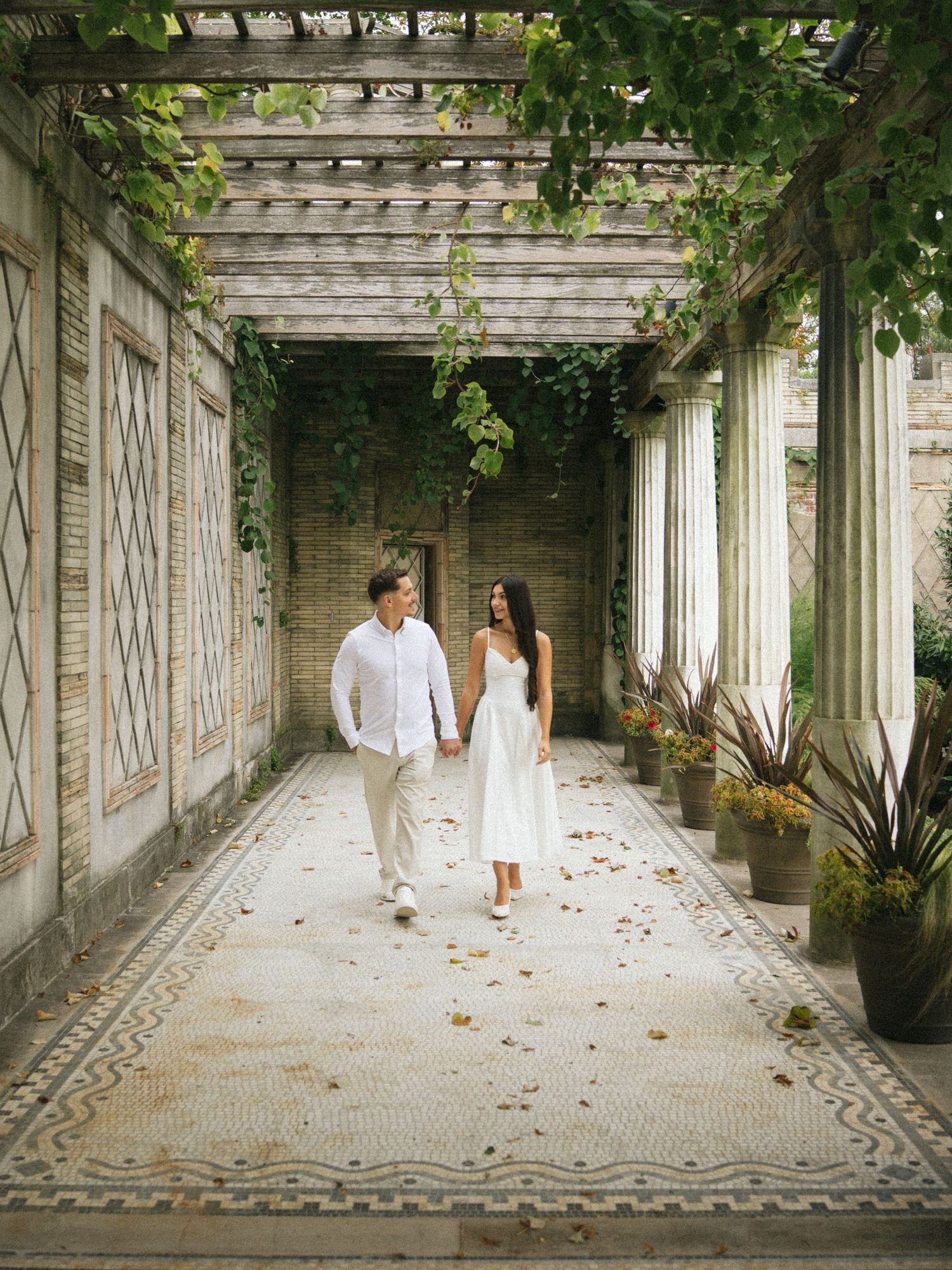 Couple in white attire walking hand-in-hand through an elegant vine-covered pergola with mosaic tile flooring.