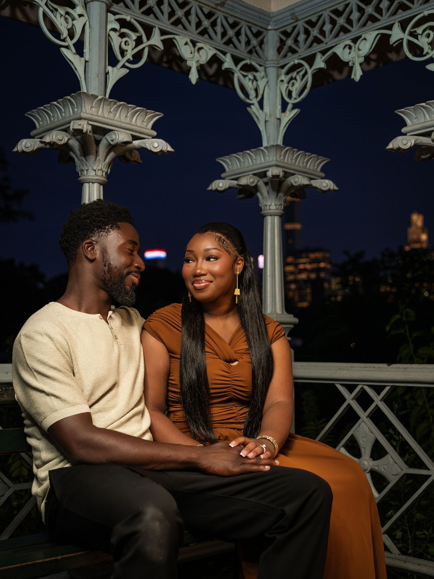 Couple sitting together at night in a decorative gazebo with city lights in the background.