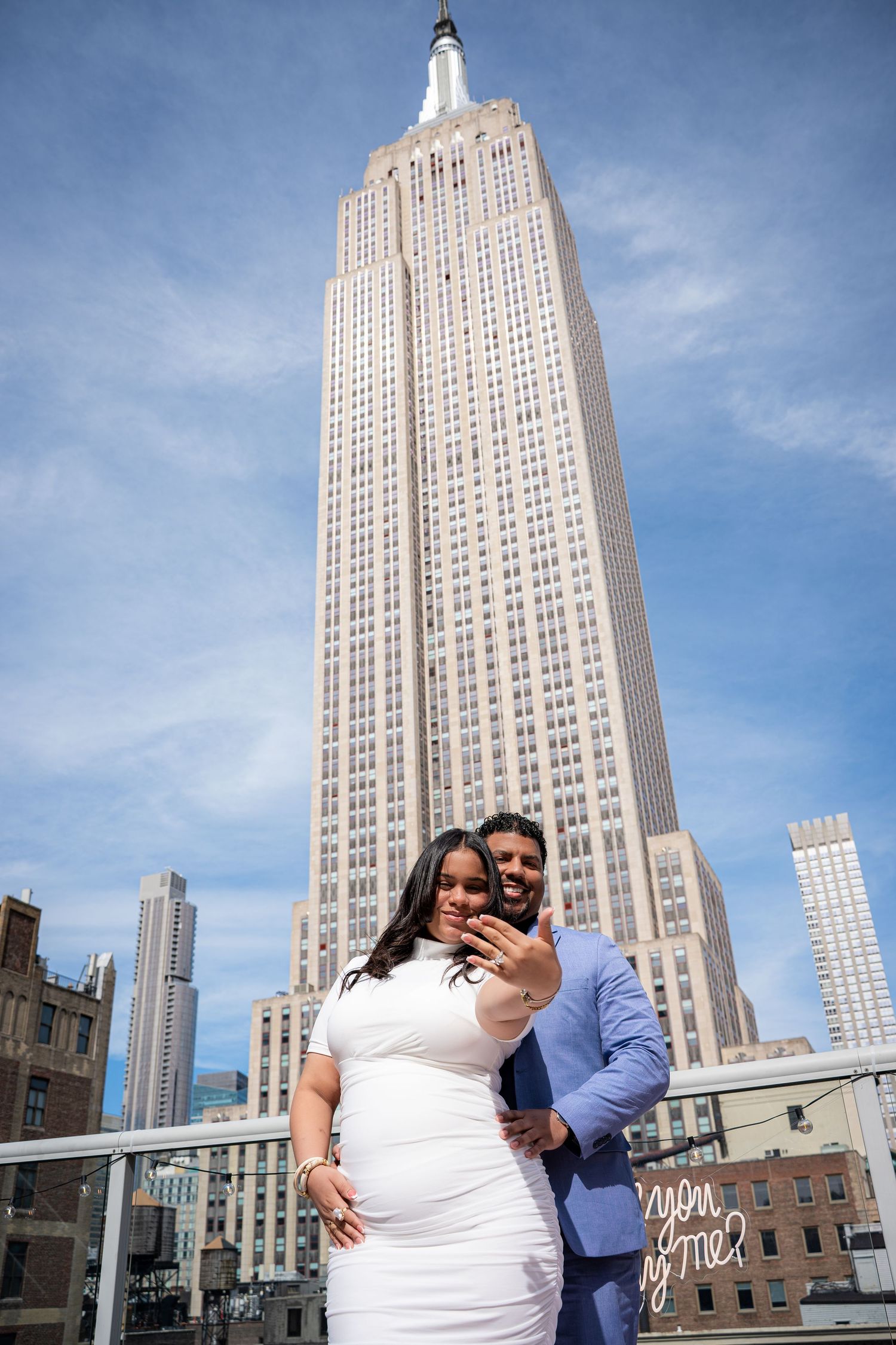 Couple takes selfie in front of the Empire State Building in NYC, woman in white dress, man in blue suit.