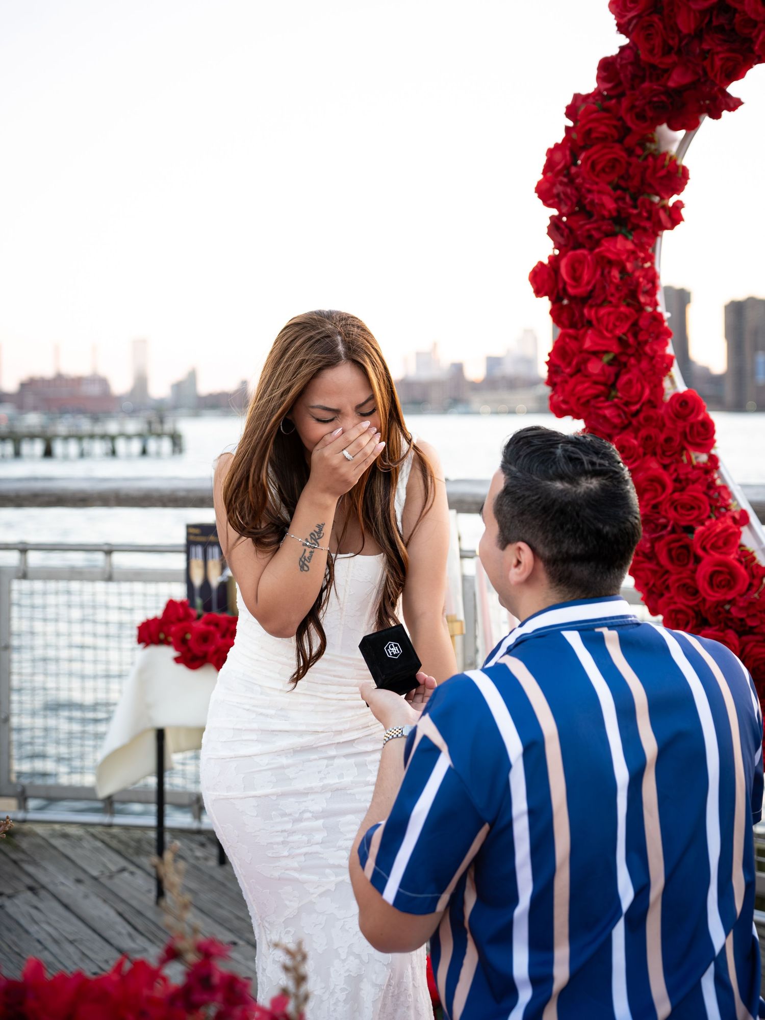 Man kneeling to propose to emotional woman in white dress beside red rose arch with NYC skyline backdrop.