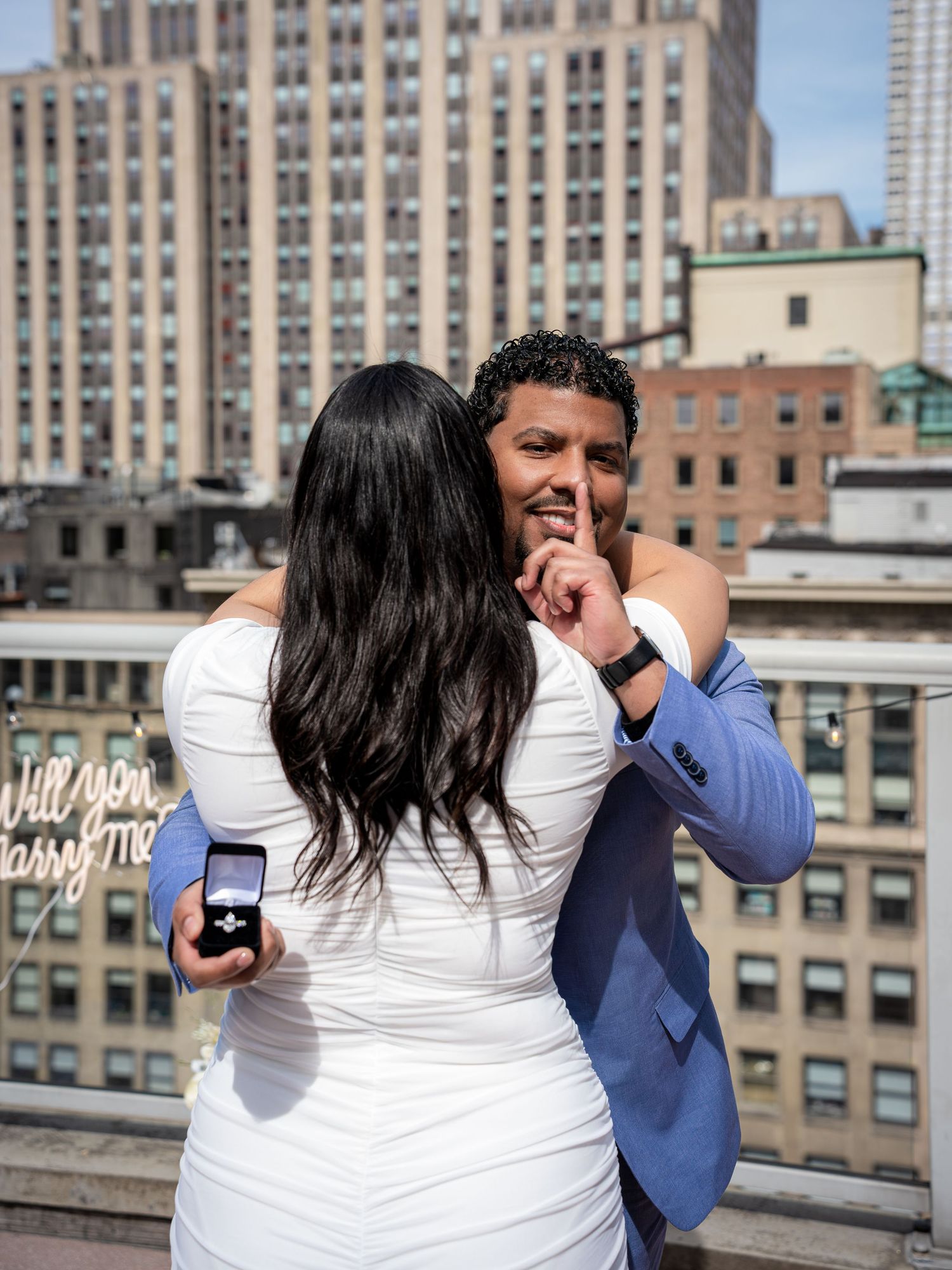 Man in blue suit holds engagement ring behind woman in white dress on NYC rooftop, gesturing shhh.