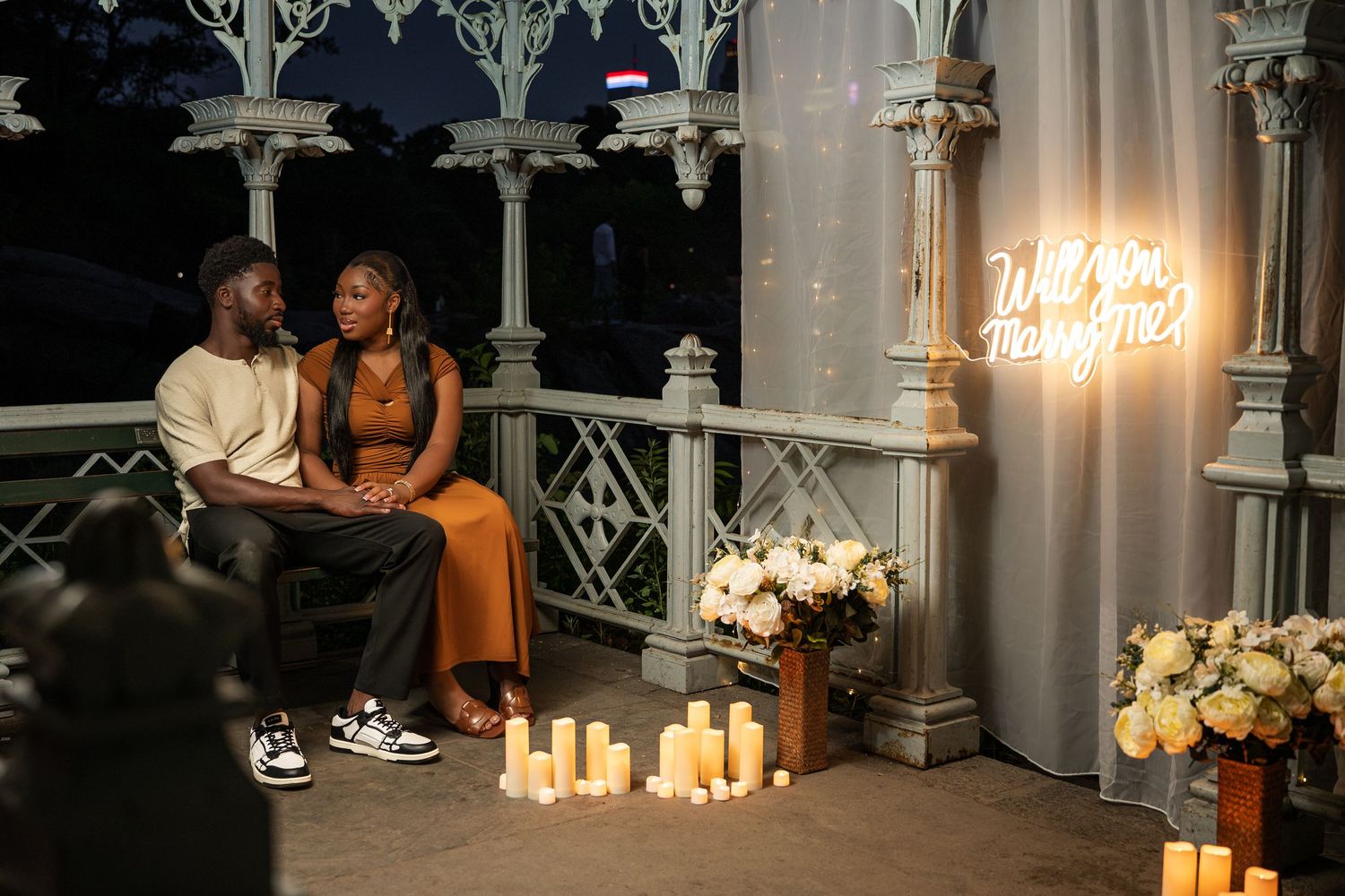 Romantic proposal setup with neon sign, candles, and flowers on a balcony at night with a couple seated together.