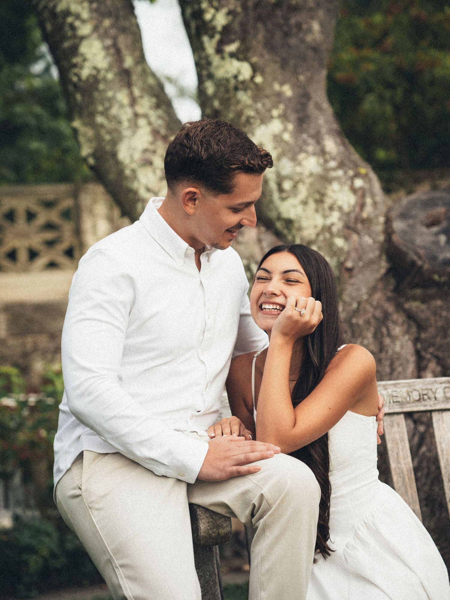 A couple in white outfits laughing together on a bench outdoors, surrounded by lush greenery.