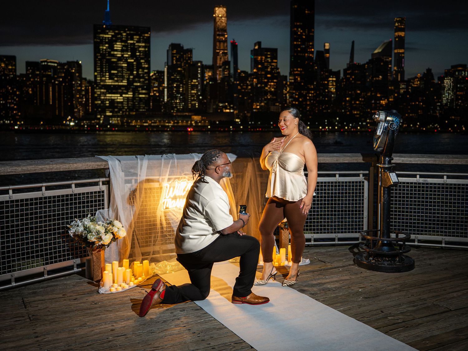 Man proposing on one knee to woman on waterfront pier with NYC skyline illuminated at night behind them.