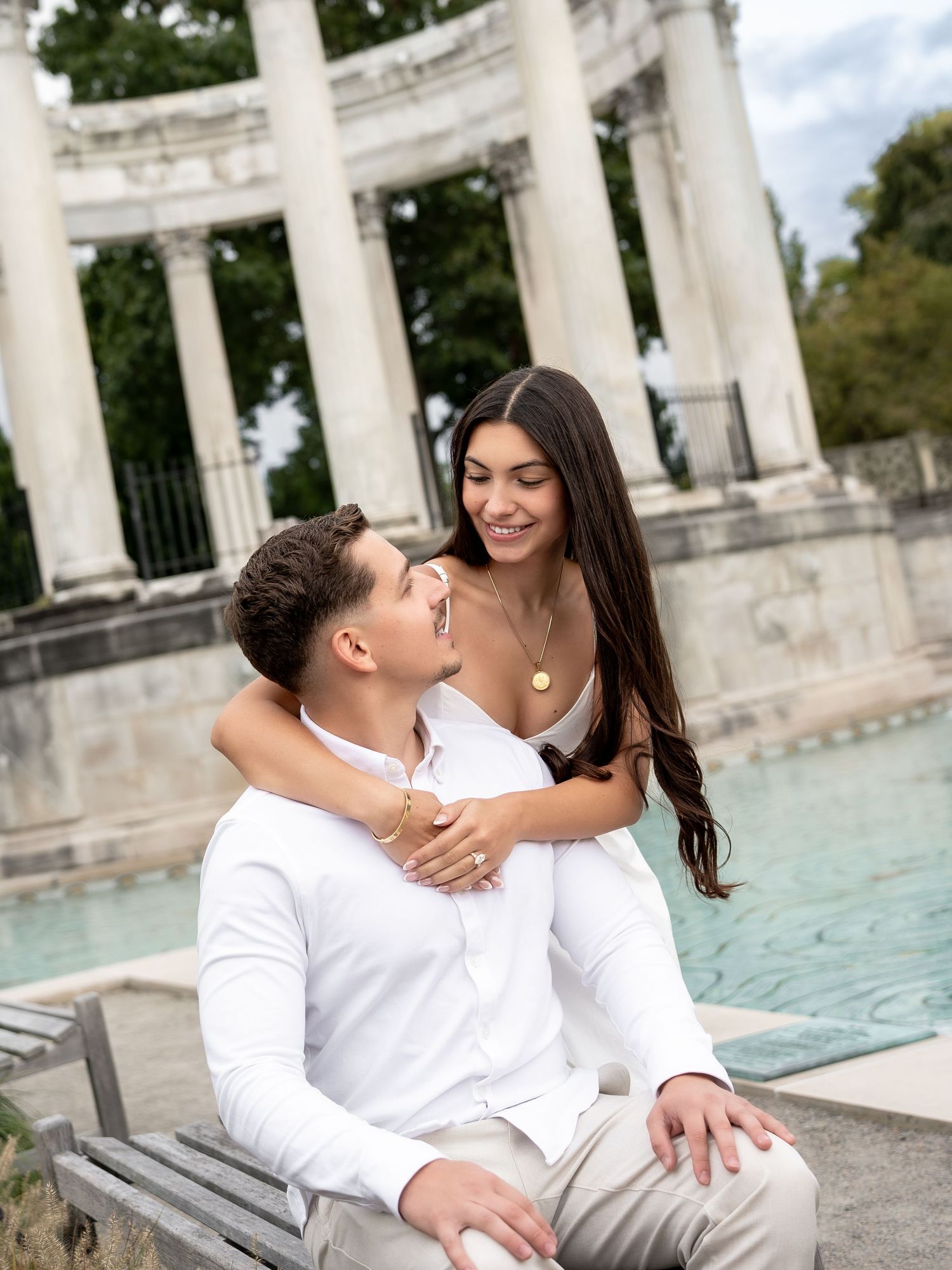 Couple in white outfits sharing a romantic moment by a fountain with classical columns in the background.