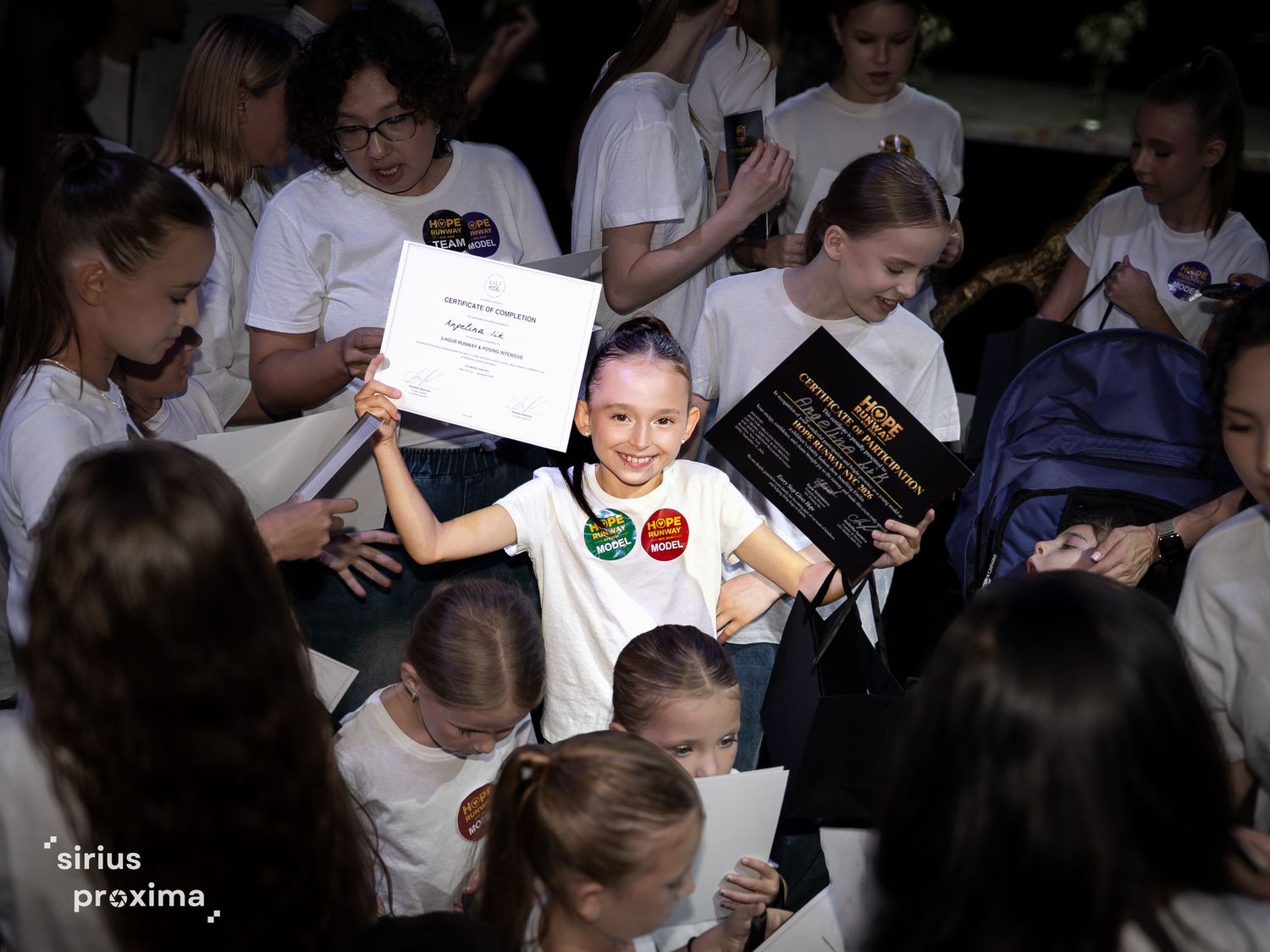 Young girl proudly holds certificate and award at school competition surrounded by peers in white t-shirts.