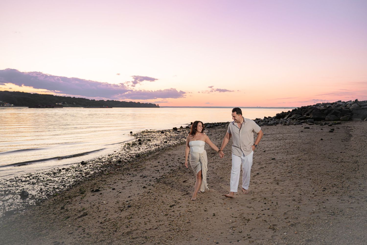 Couple holding hands walking on sandy beach at sunset with pink and purple sky reflecting on calm water.