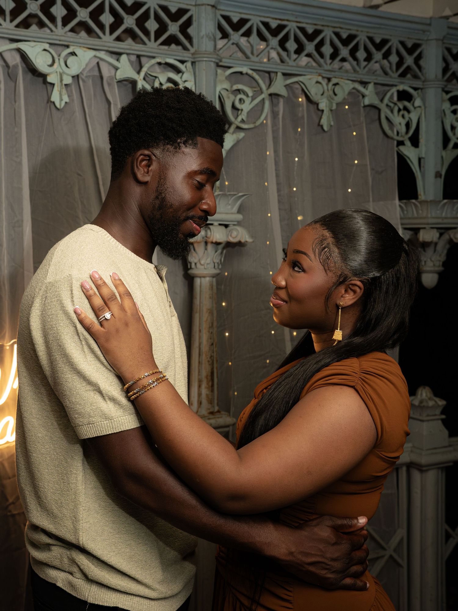 A couple shares a romantic moment, gazing into each other's eyes, dressed in casual earth tones against a decorative backdrop.