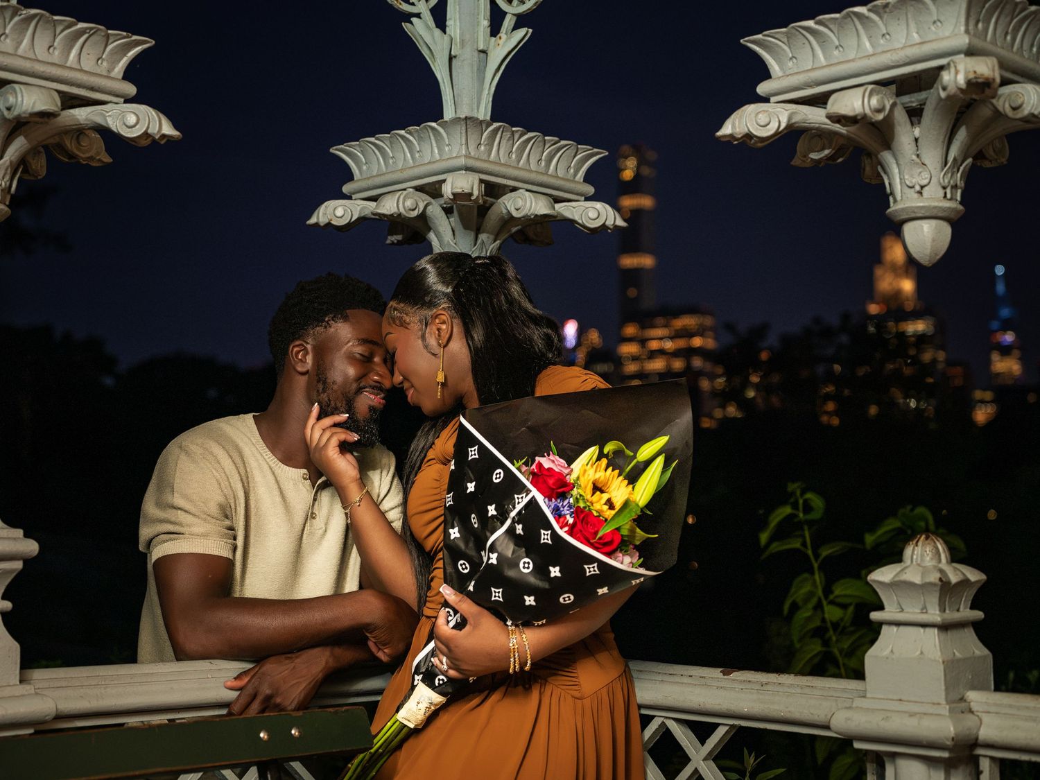 Couple sharing a romantic moment on a bridge at night with city skyline backdrop, woman holding colorful bouquet.