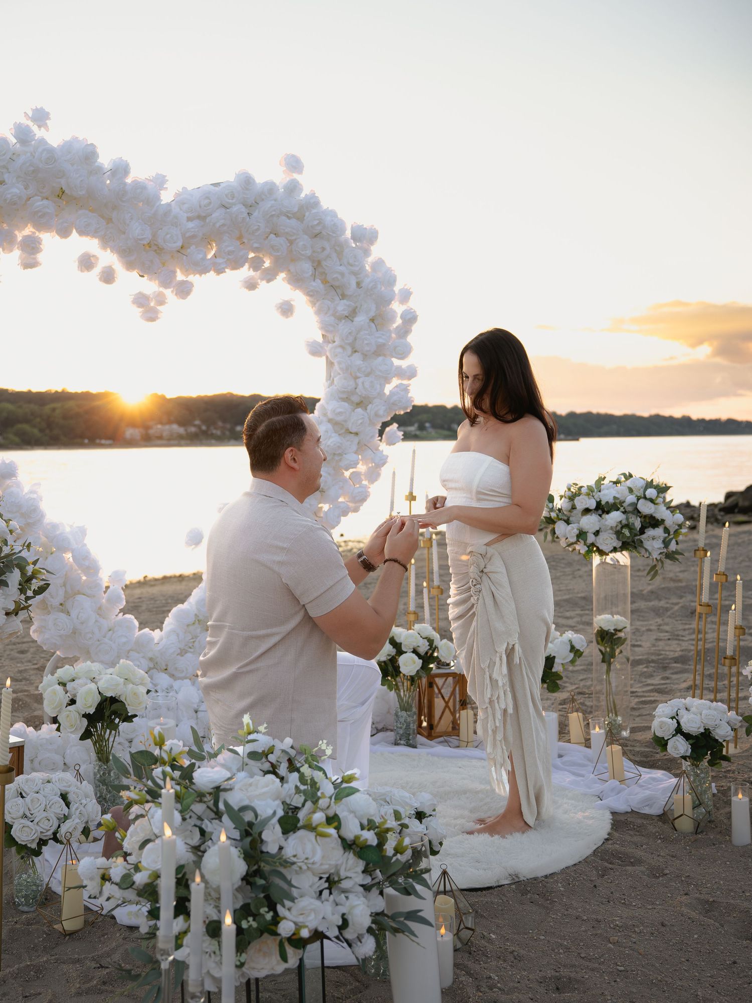 Man proposing to woman on beach at sunset with white floral heart arch and candles.