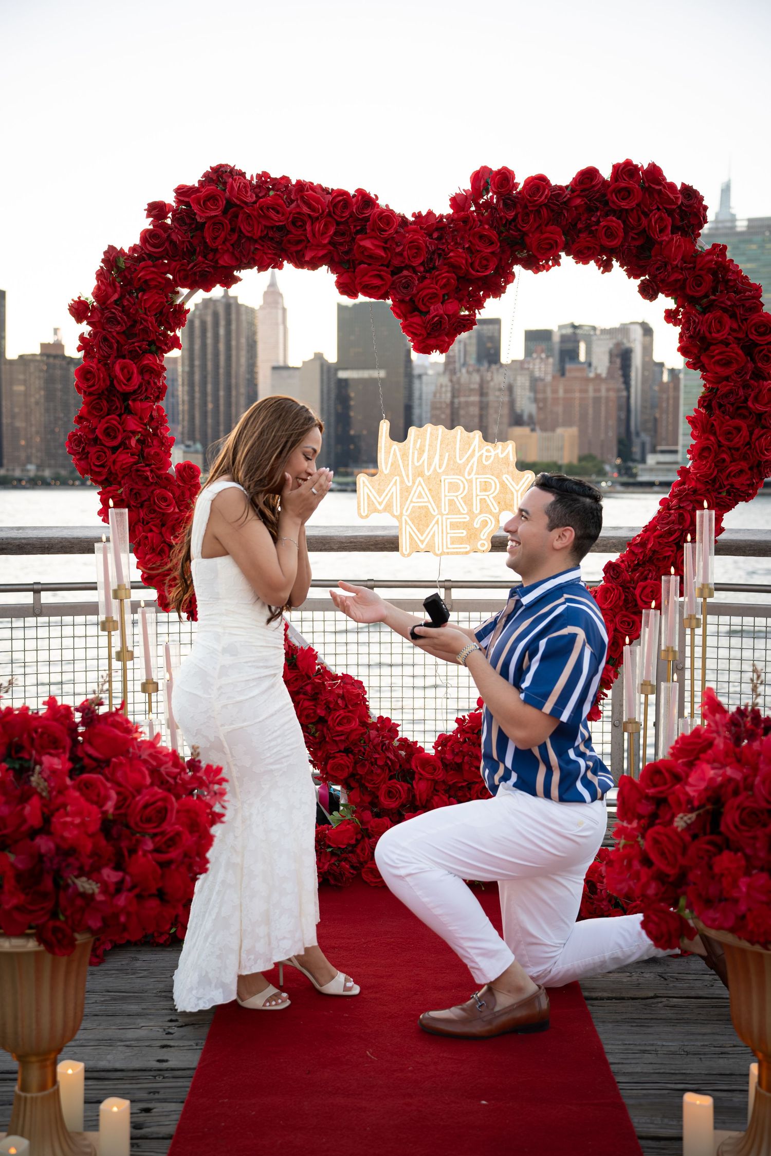 Man proposes to woman on one knee under red rose heart arch with NYC skyline backdrop and Will You Marry Me sign.