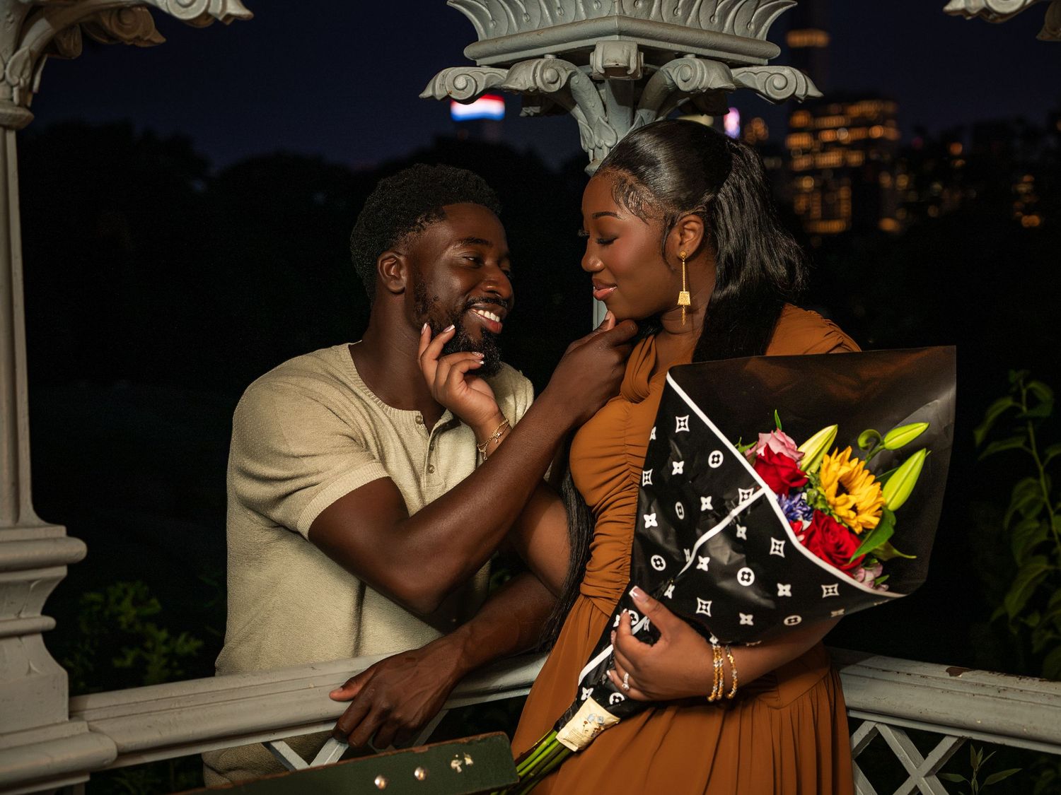 Couple sharing a romantic moment at night on a bridge, woman holding a Louis Vuitton bouquet of sunflowers.