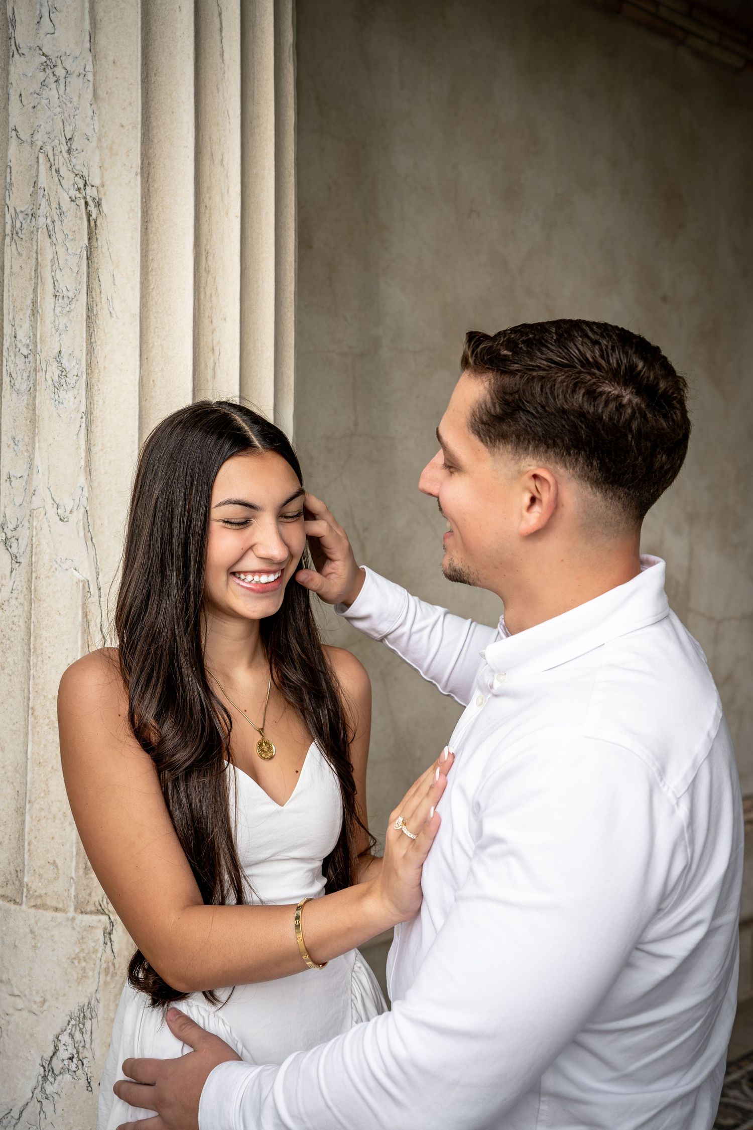 Couple in white outfits sharing a tender moment by marble columns, man gently touching woman's hair as she smiles.