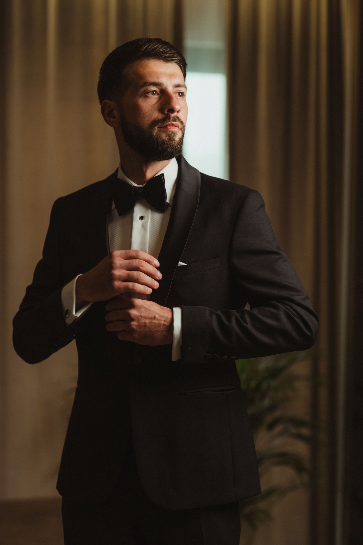 Elegant bearded groom in black tuxedo with bow tie buttoning jacket near curtained window.