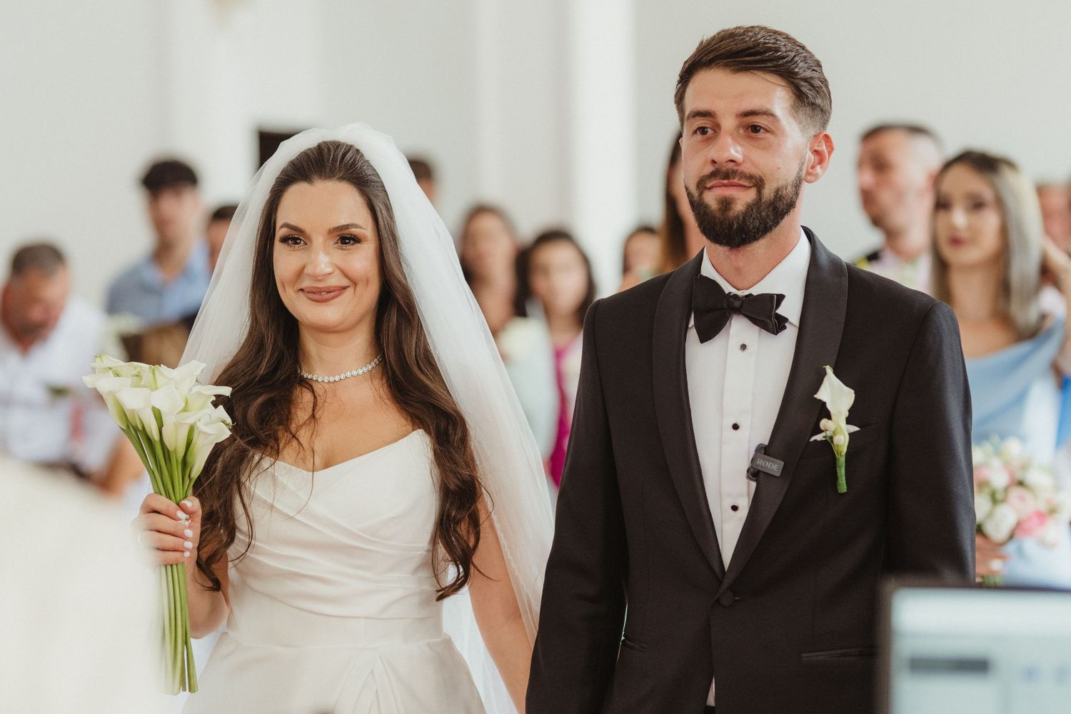 Bride and groom walking down church aisle, bride holding white calla lilies, guests seated behind.