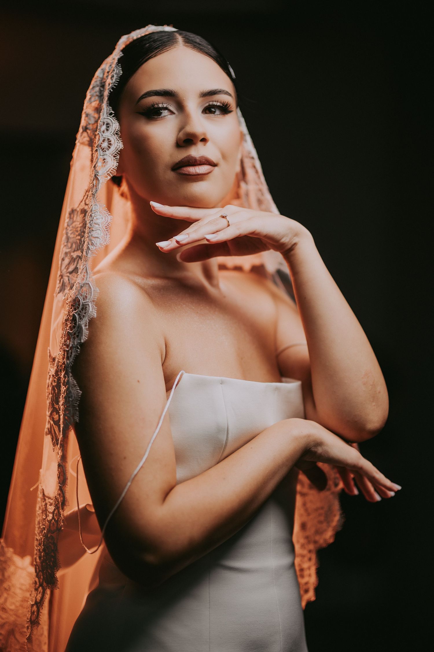Elegant bride in strapless gown wearing intricate lace mantilla veil posing gracefully in warm golden light.