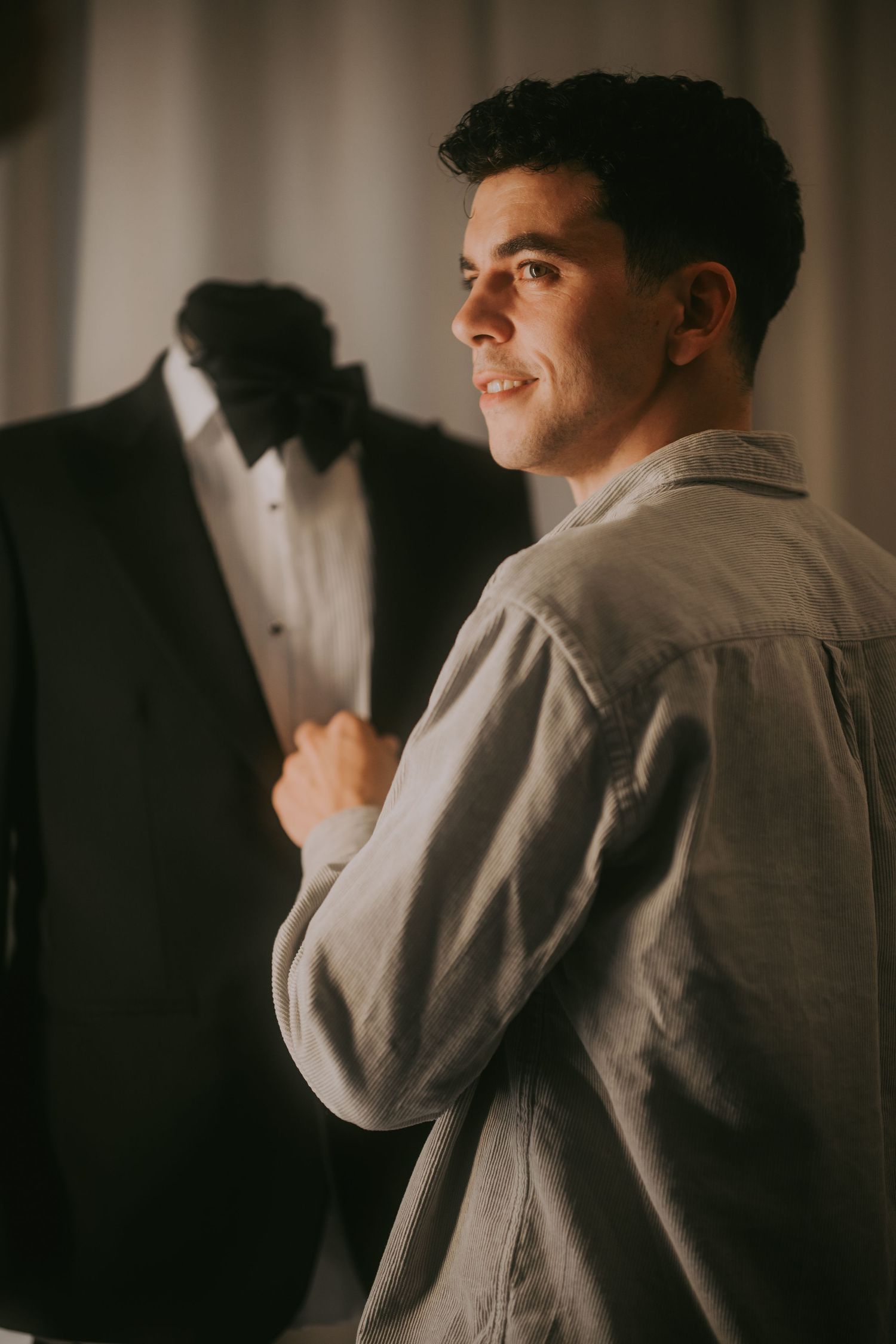 Smiling groom in gray shirt admiring a black tuxedo with bow tie on a dress form. Epicu Fotograf Drobeta-Turnu Severin