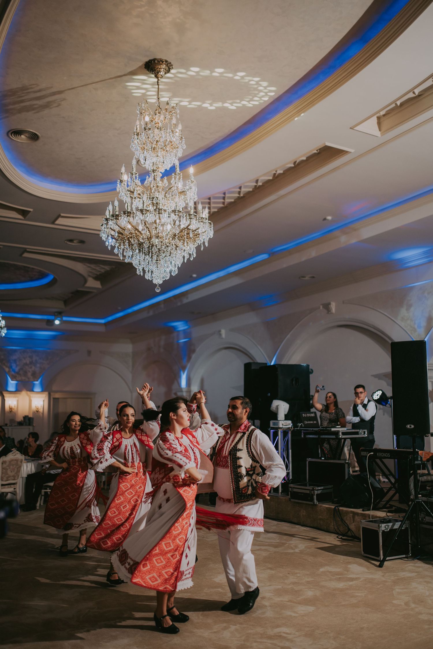 Traditional folk dancers in embroidered costumes performing under a crystal chandelier at a wedding reception. Epicu Fotograf Drobeta-Turnu