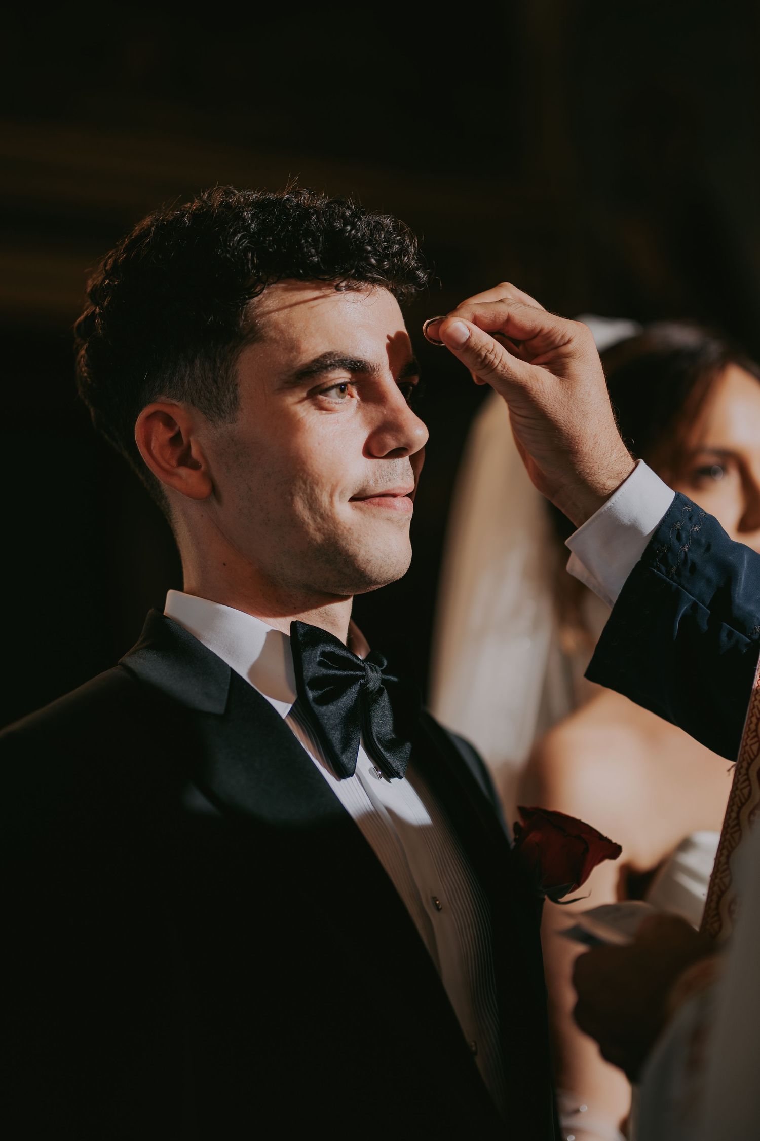 Groom in black tuxedo smiles as officiant performs anointing ritual during Orthodox wedding ceremony. Epicu Fotograf Drobeta-Turnu Severin