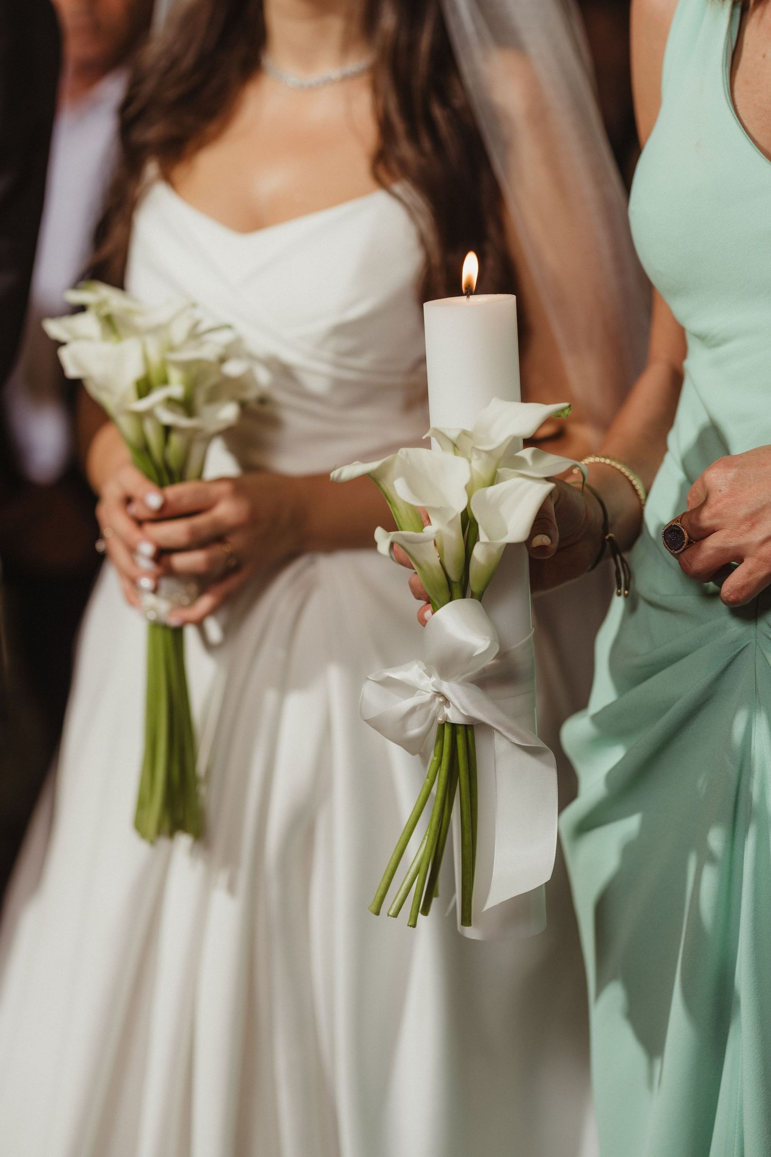 Bride in white gown holds calla lily bouquet and lit candle beside bridesmaid in mint dress during wedding ceremony.