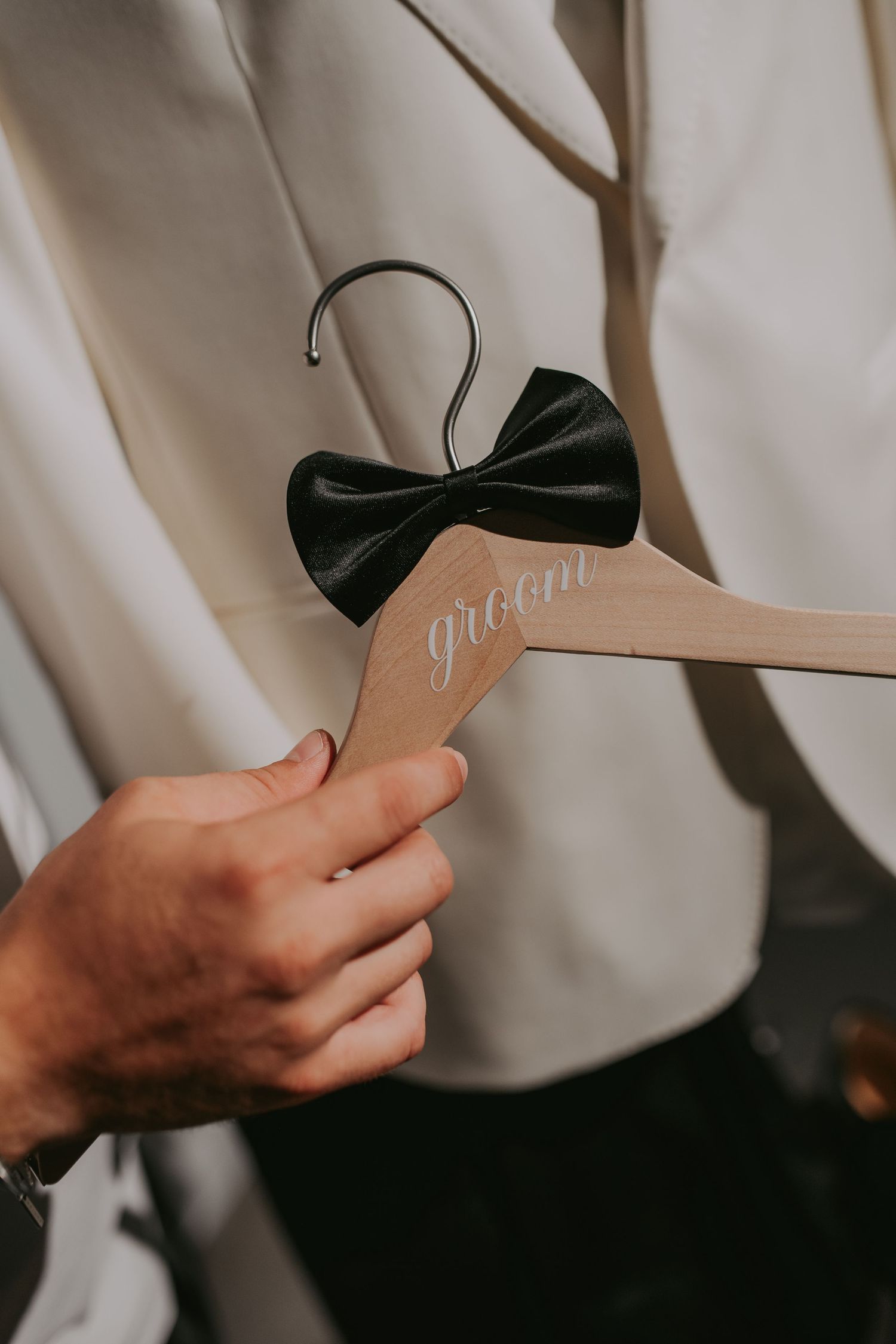Hand holding a wooden groom hanger with black bow tie against a white wedding suit. Epicu Fotograf Drobeta-Turnu Severin