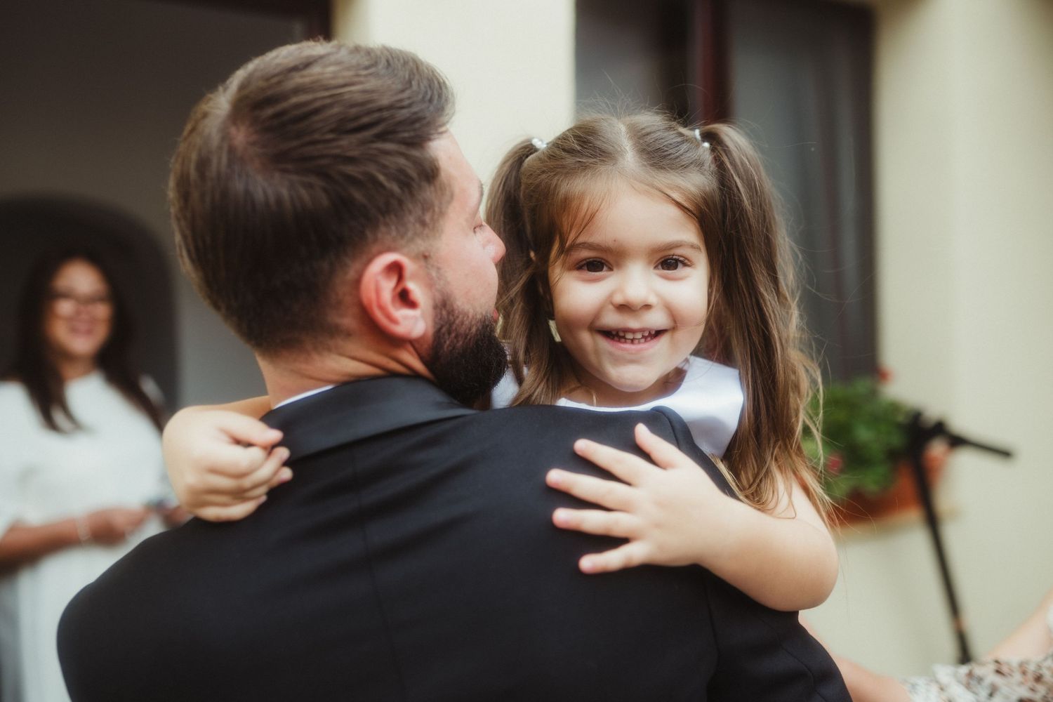 Groom in black tuxedo holding smiling little flower girl at outdoor wedding ceremony.