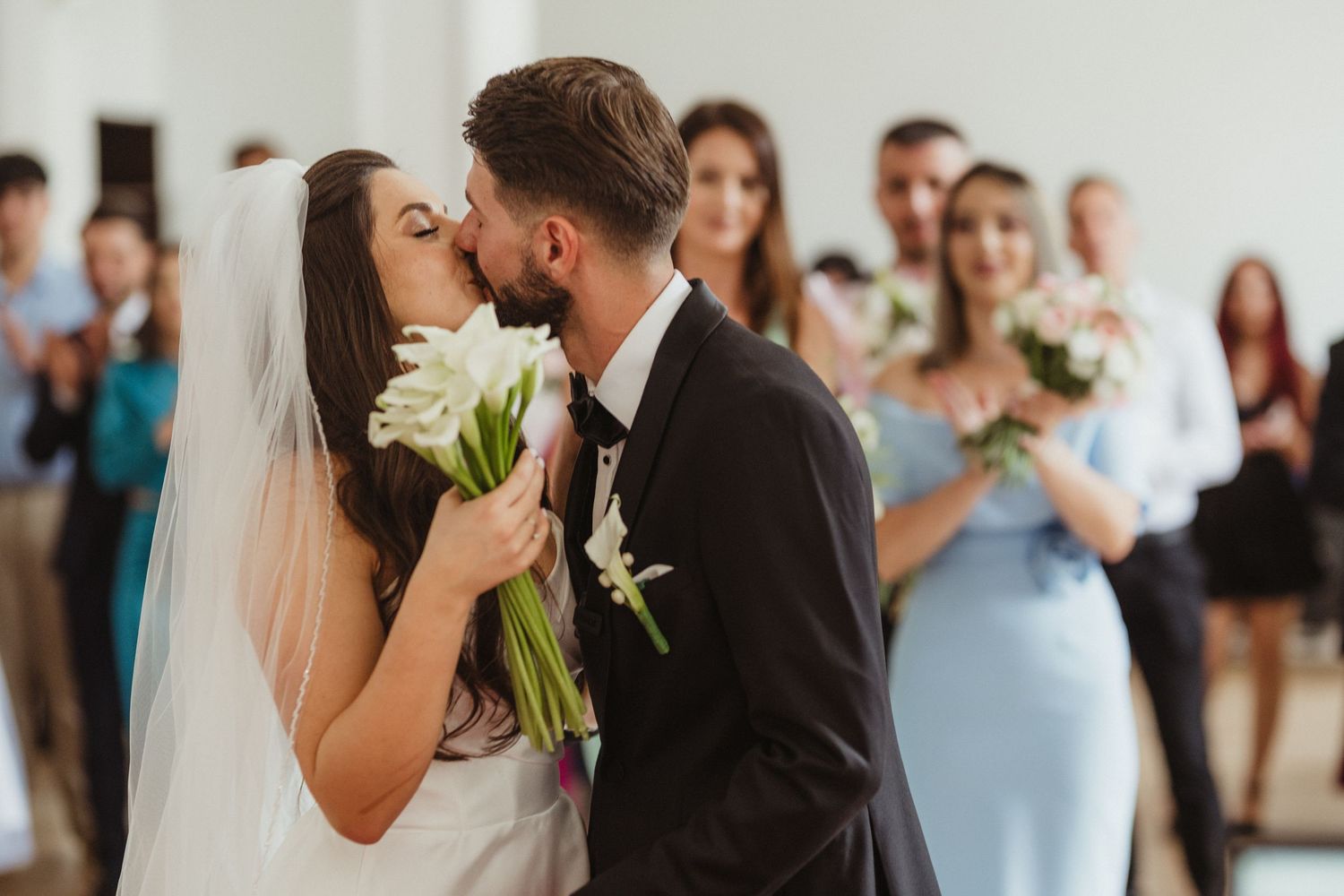 Bride and groom share first kiss holding white calla lily bouquet, guests applauding behind them.