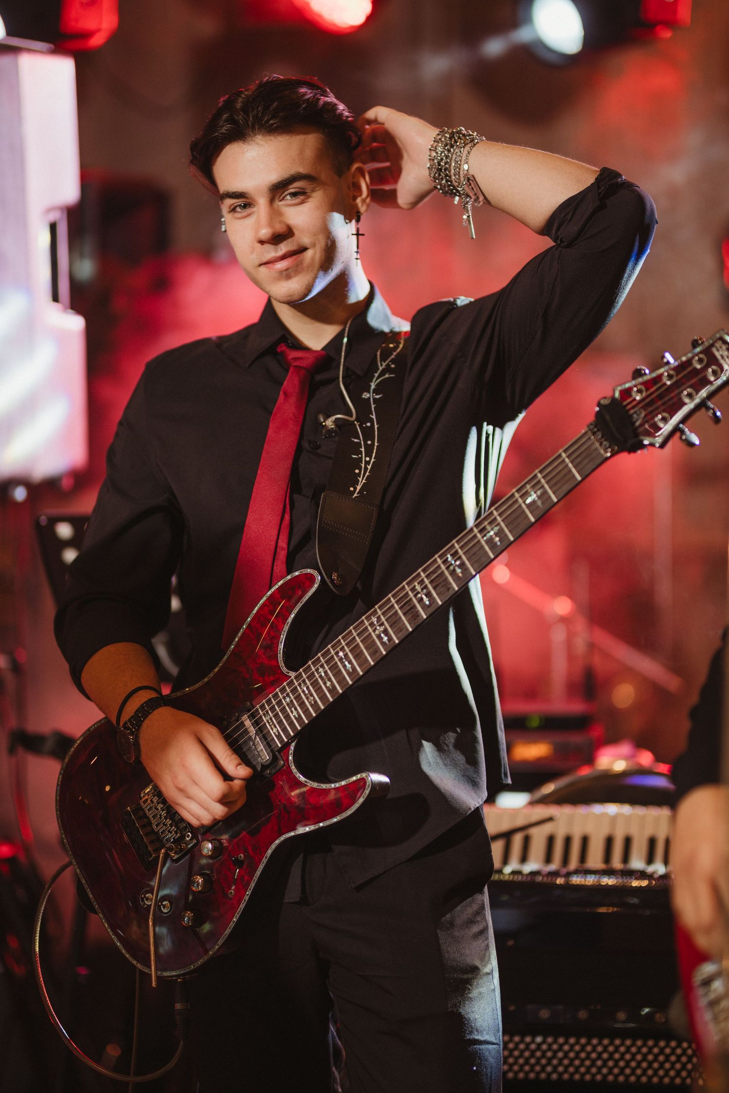 Young male guitarist in black shirt and red tie posing with electric guitar on stage.