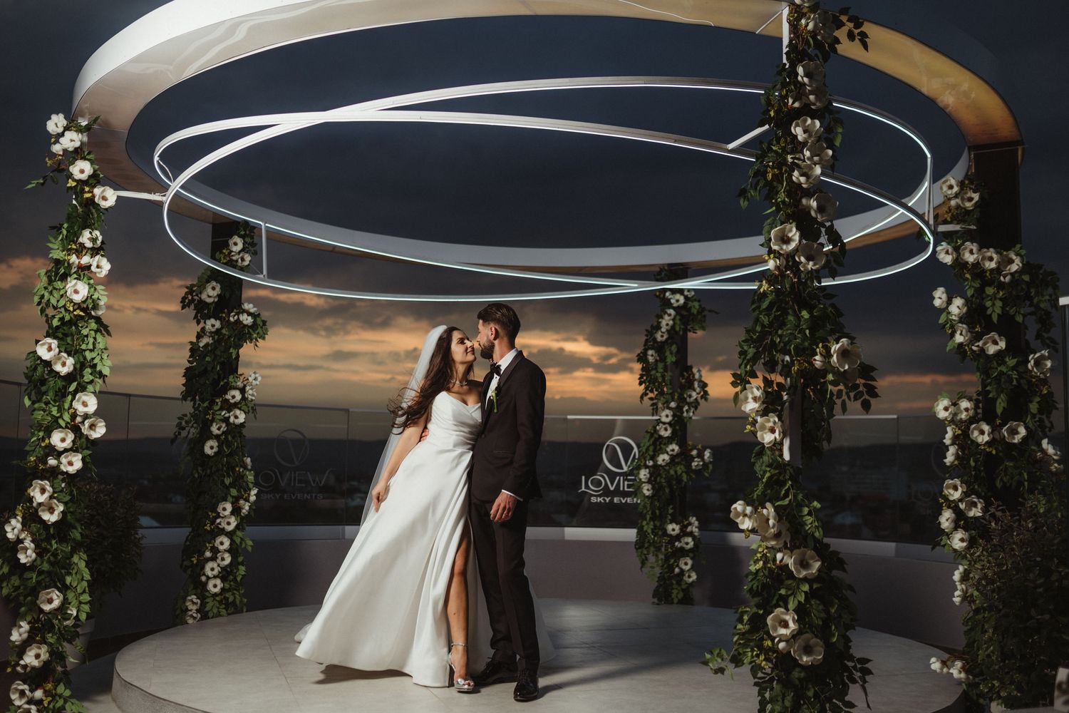 Newlyweds share a kiss under a circular floral arch at a rooftop wedding venue at dusk.