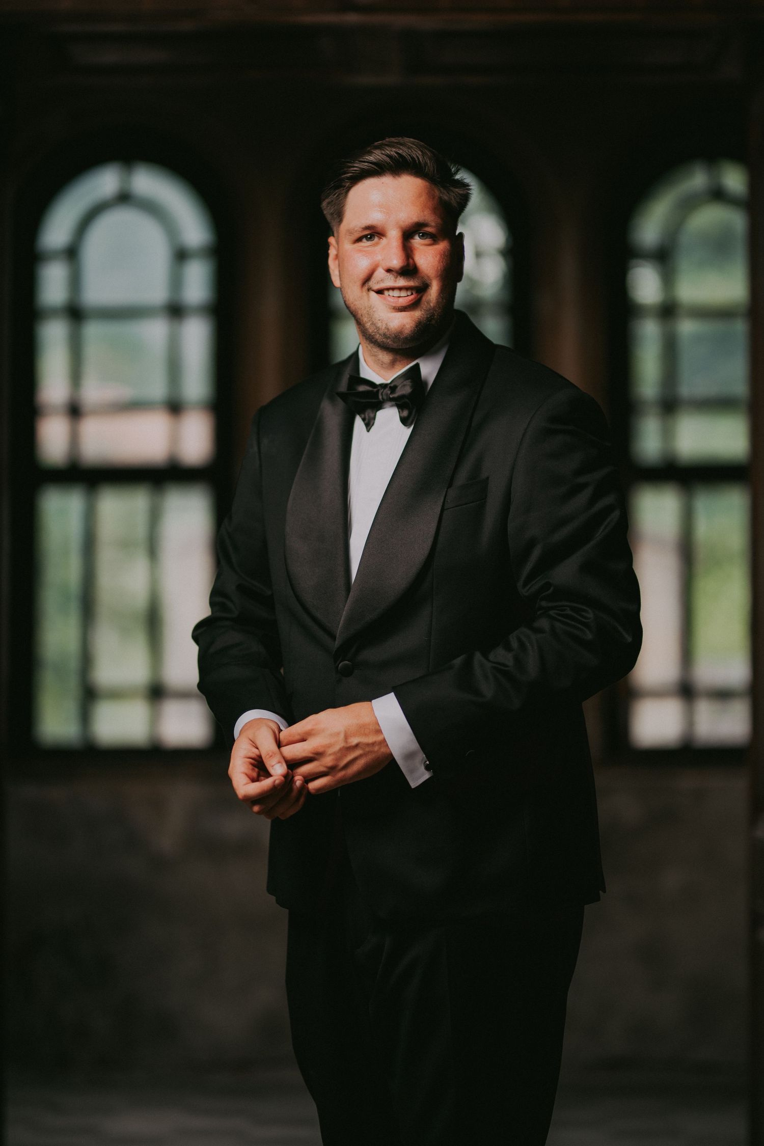 Handsome groom in black tuxedo and bow tie smiling confidently in a grand historic venue.