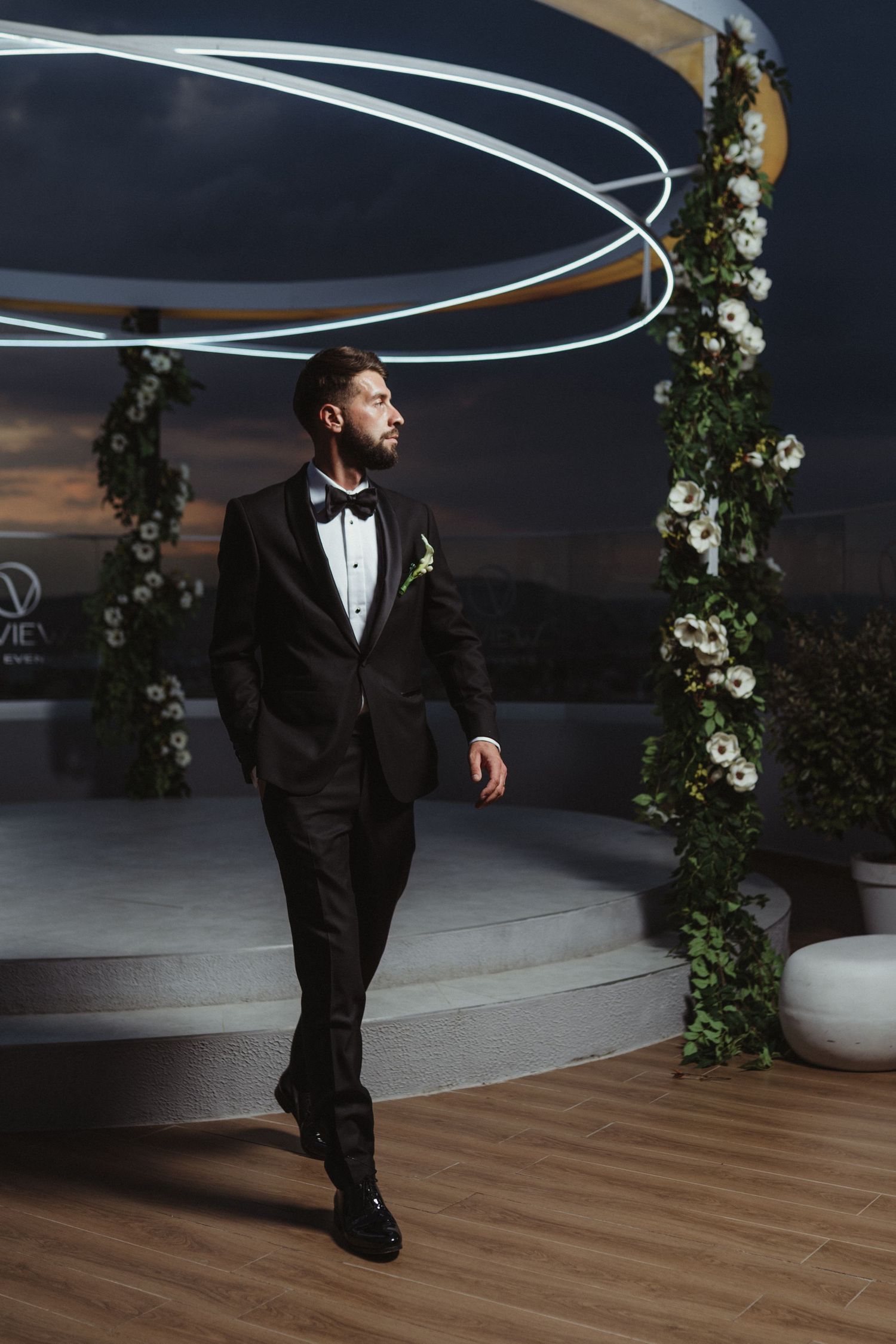 Dapper groom in black tuxedo with bow tie stands confidently at rooftop wedding venue with floral arch at dusk.