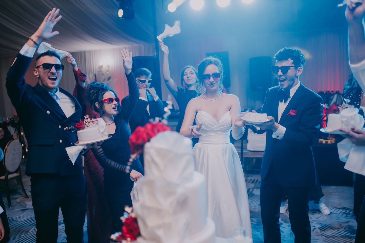 Wedding party wearing sunglasses celebrating around a white tiered cake at a festive reception. Epicu Fotograf Drobeta-Turnu Severin