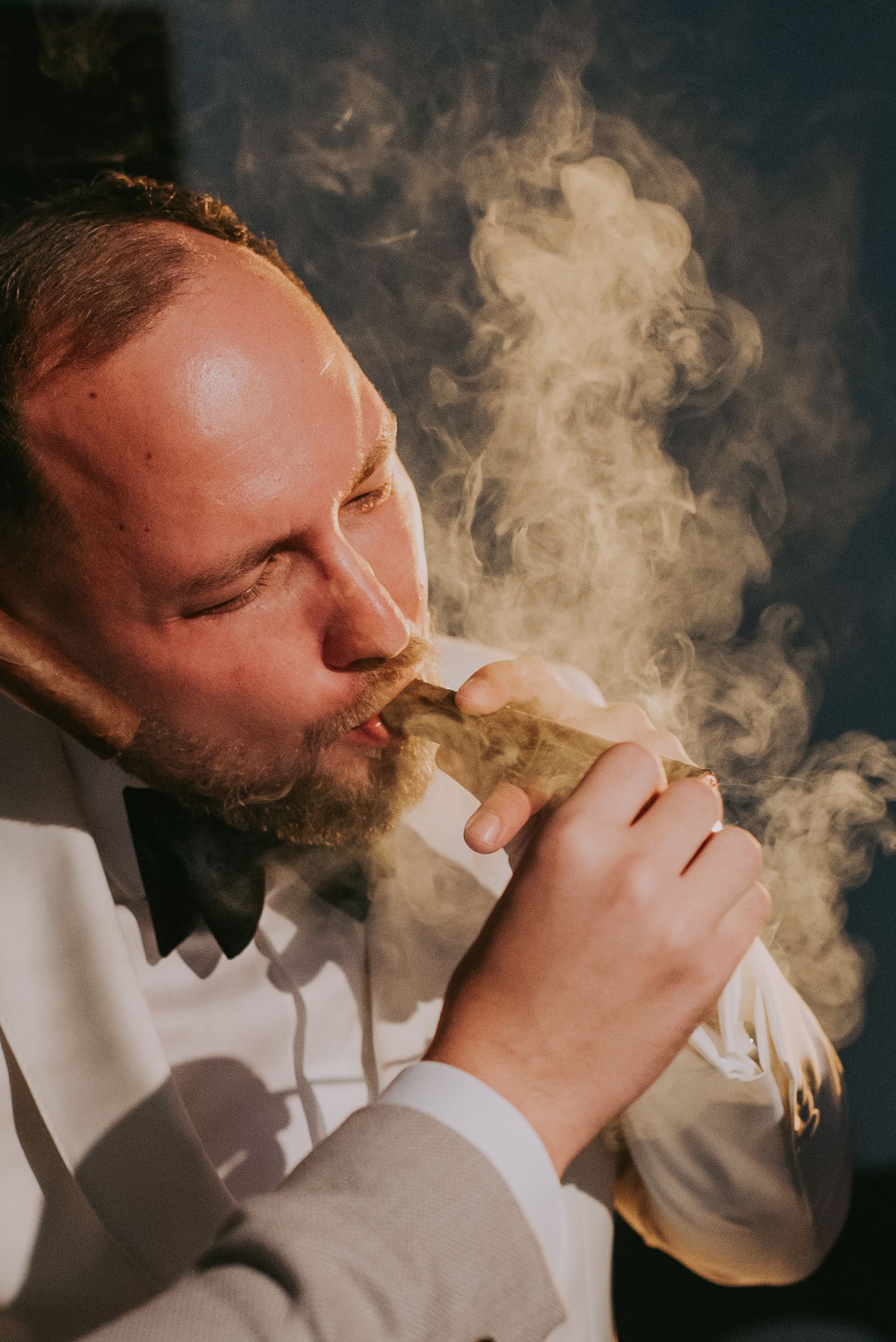 Groom in white tuxedo smoking a cigar with dramatic smoke clouds surrounding his face. Epicu Fotograf Drobeta-Turnu Severin