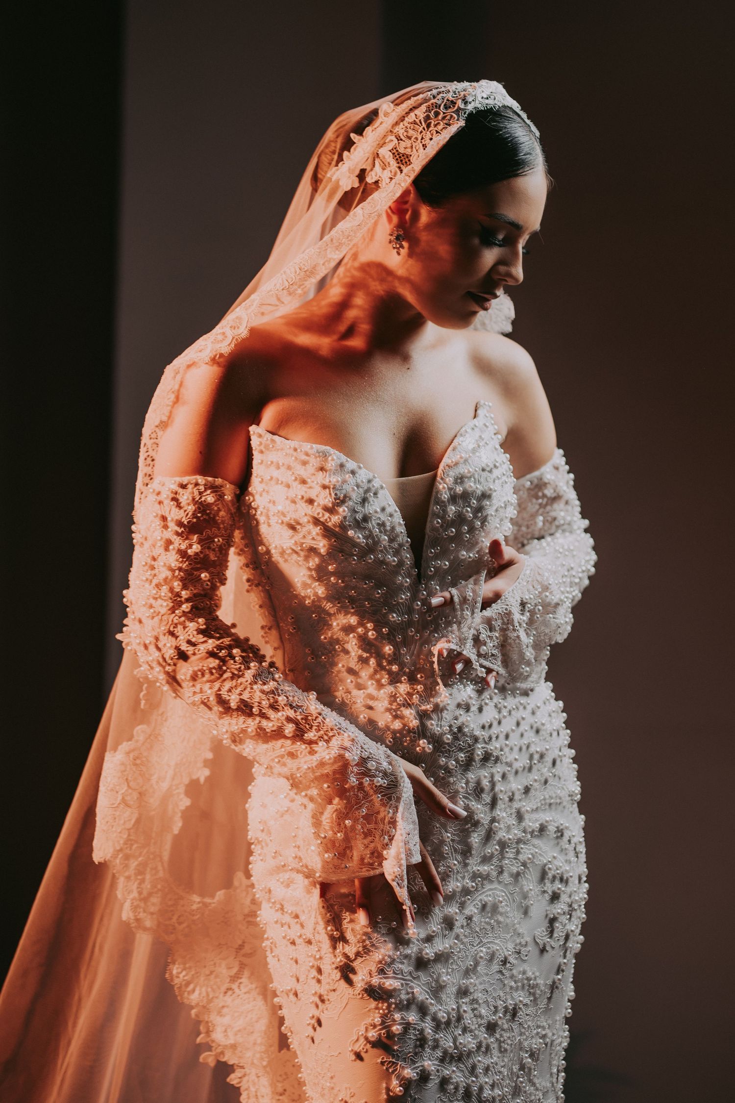 Bride in pearl-embellished off-shoulder wedding gown with flowing veil looks down.